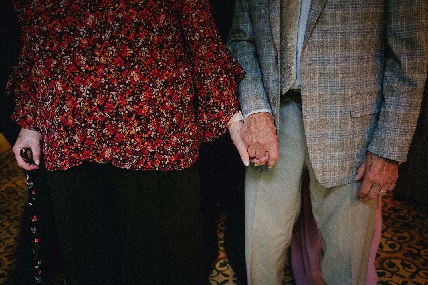 Elderly couple holding hands tenderly, woman in floral top and black skirt, man in plaid blazer and tan pants.