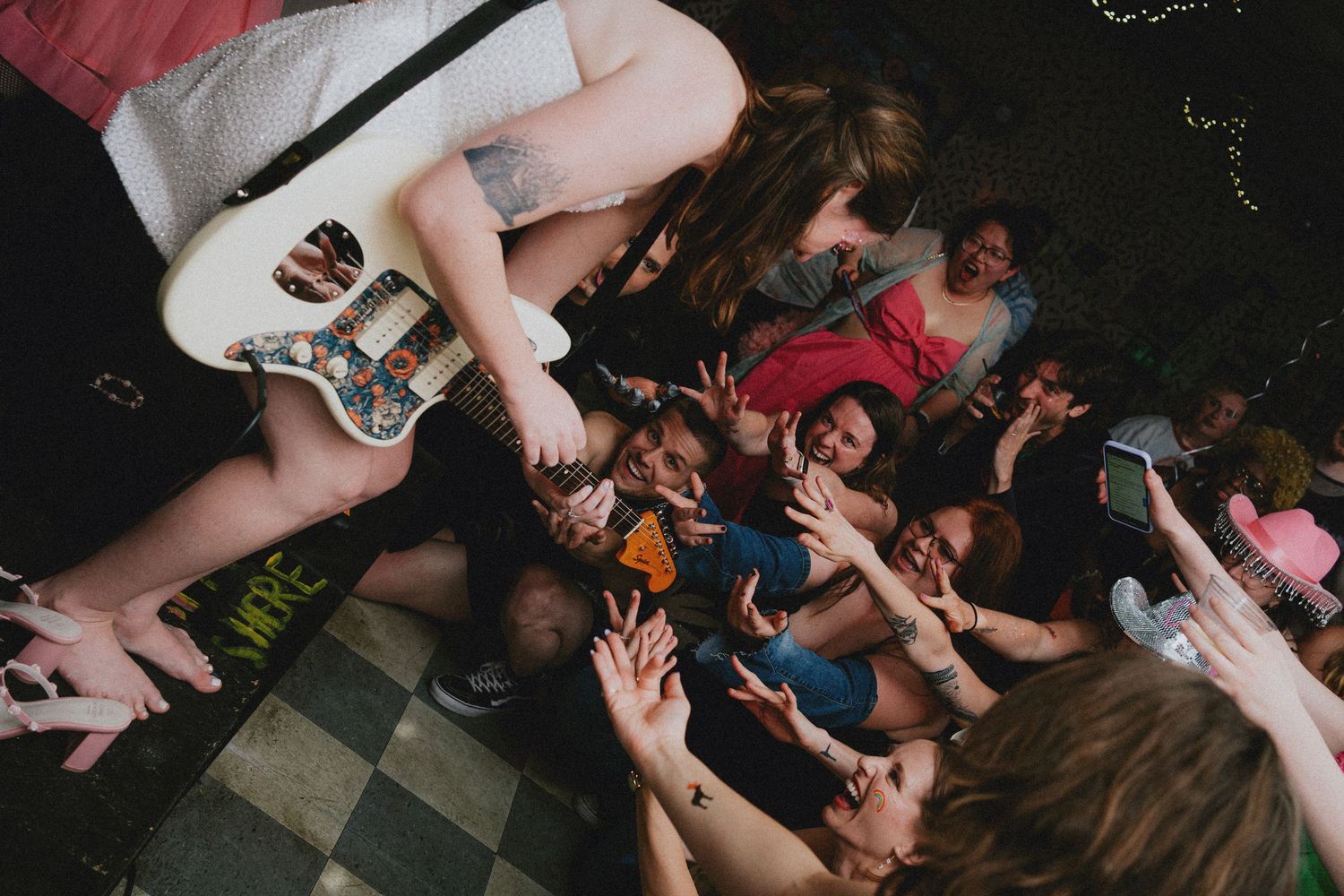 Female guitarist performs on stage while crowd reaches up toward her during an energetic live music concert.