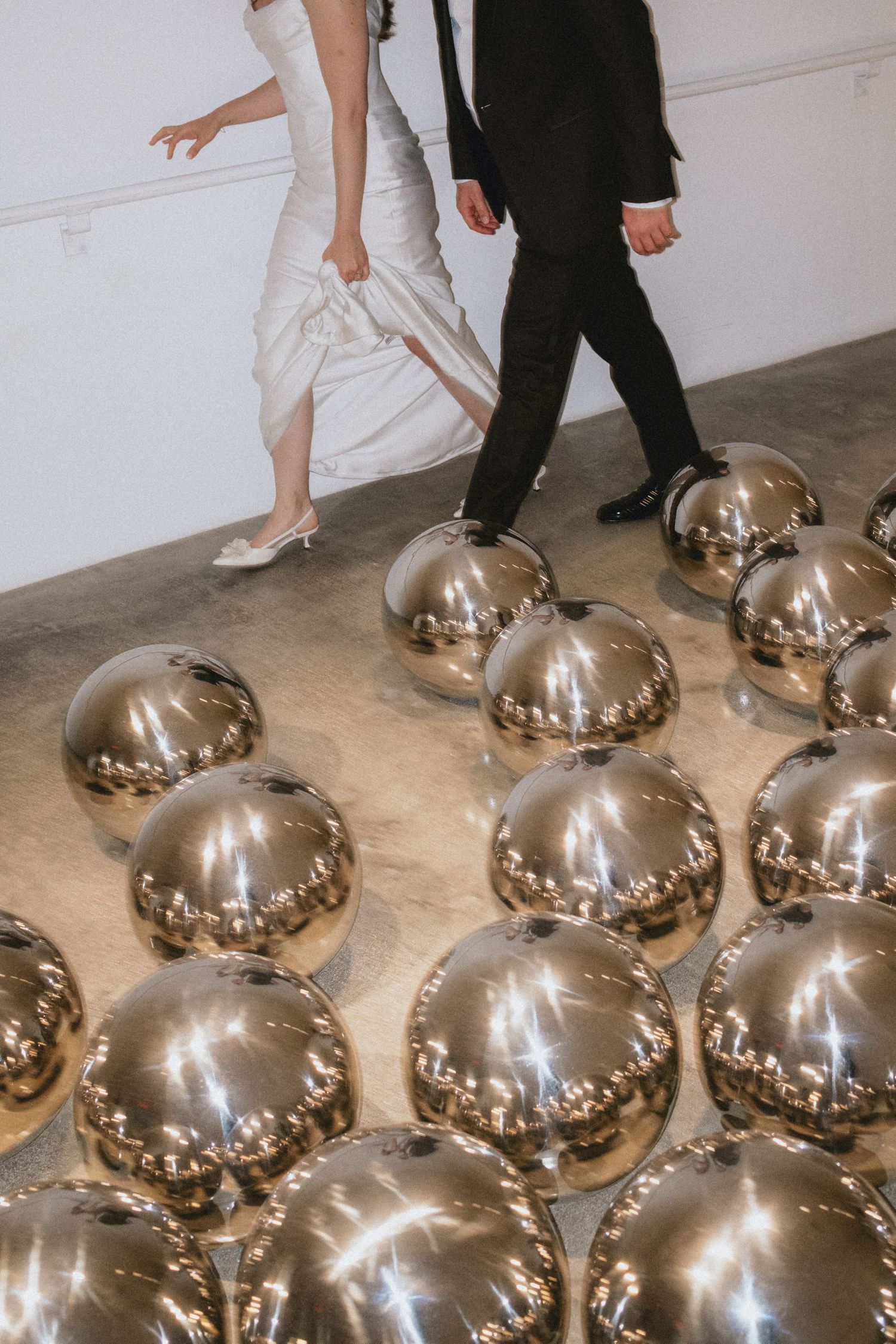 A bride in white dress and groom in black suit walk past shiny chrome metallic spheres on a gallery floor.