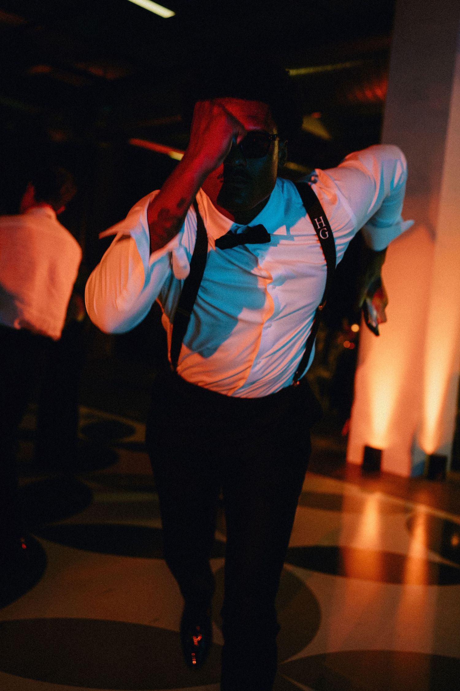 Man in white shirt, suspenders, and bow tie dancing energetically under colorful event lighting.