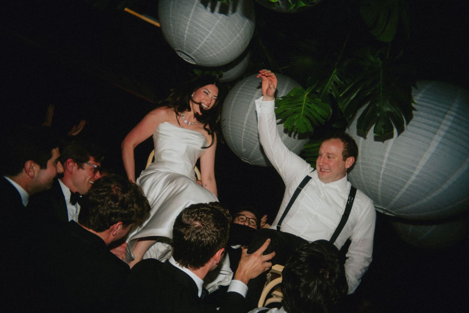 Bride and groom lifted by guests during joyful wedding reception dance under white paper lanterns.