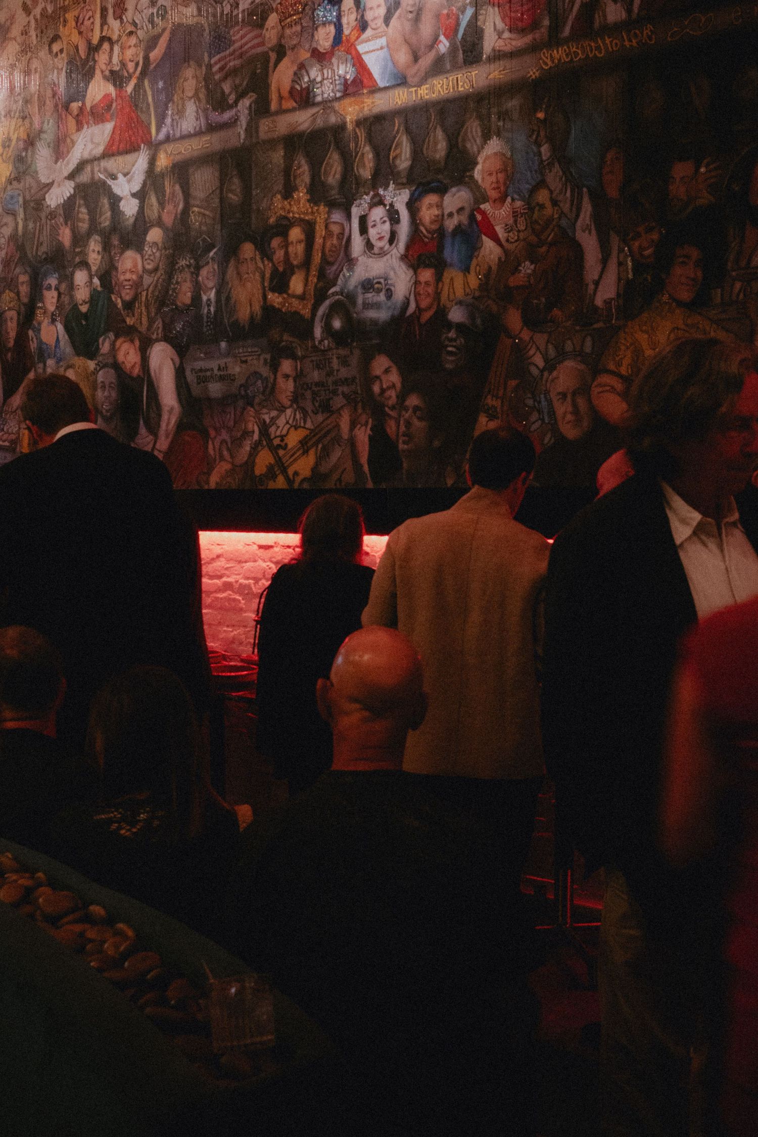 People gathered in a dimly lit bar with an elaborate colorful mural covering the wall behind them.