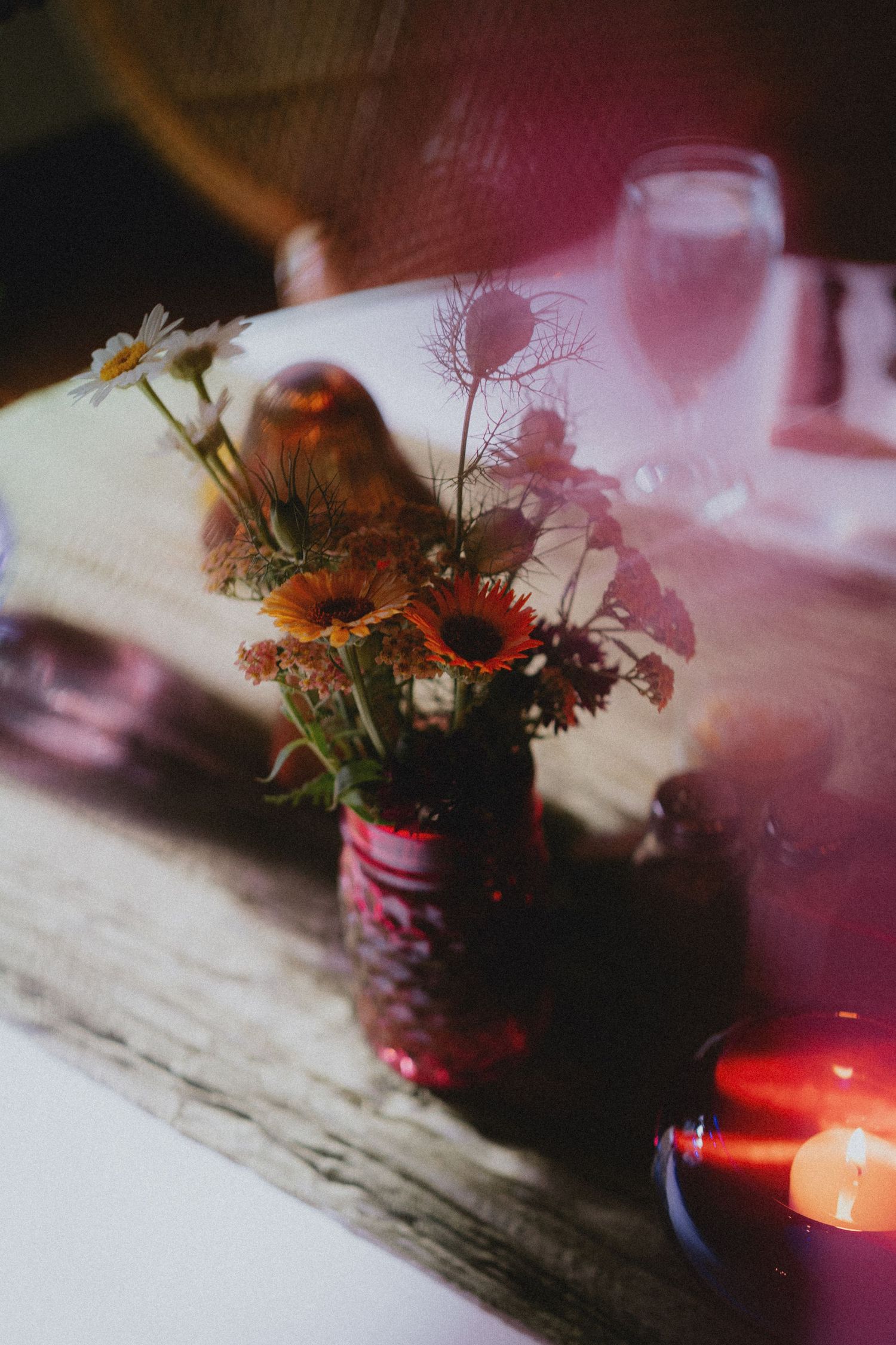 A rustic table setting with a red vase of wildflowers, candle, and champagne glass in warm, moody lighting.