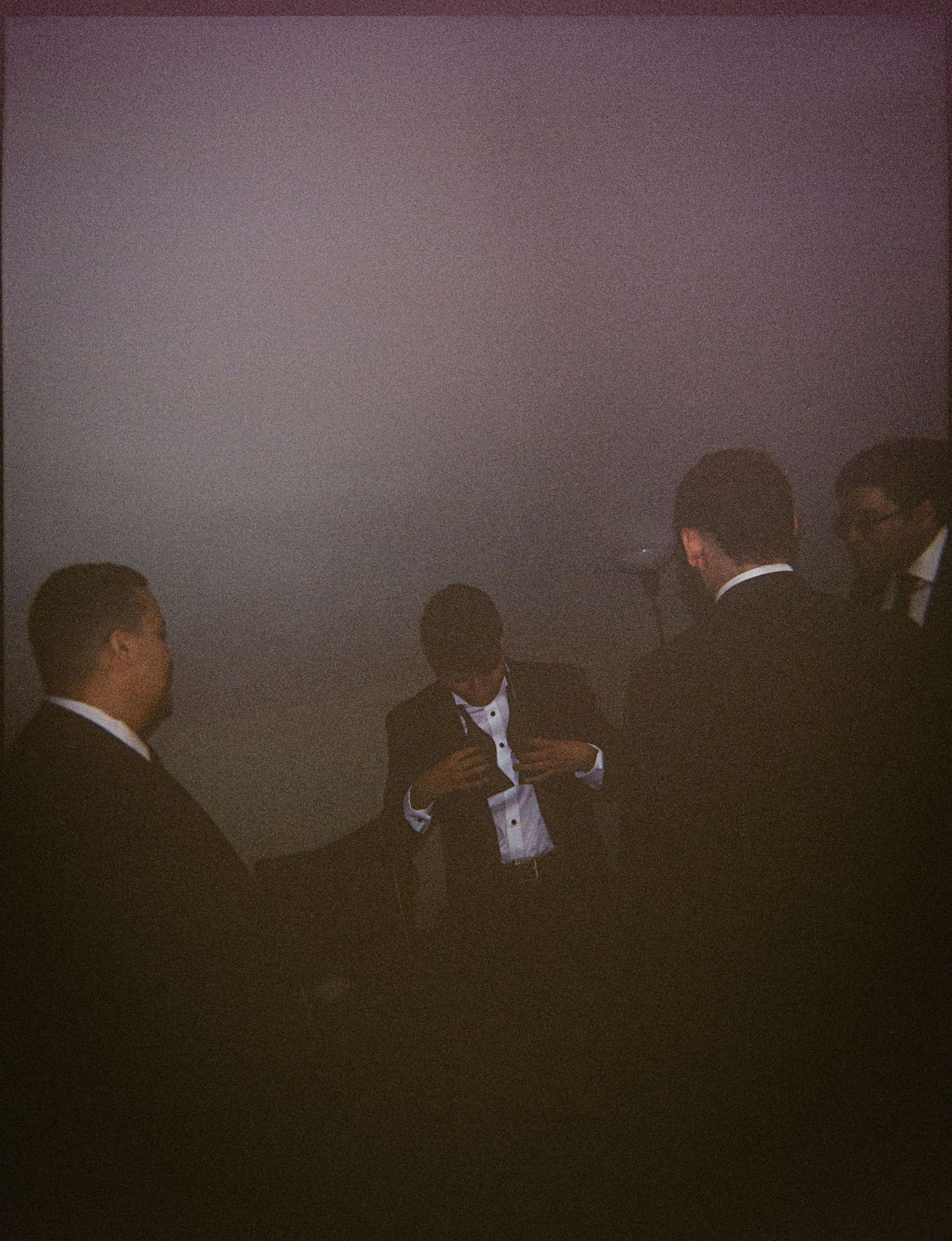 Men in formal black suits gathered together in a dimly lit, foggy venue at an upscale evening event.