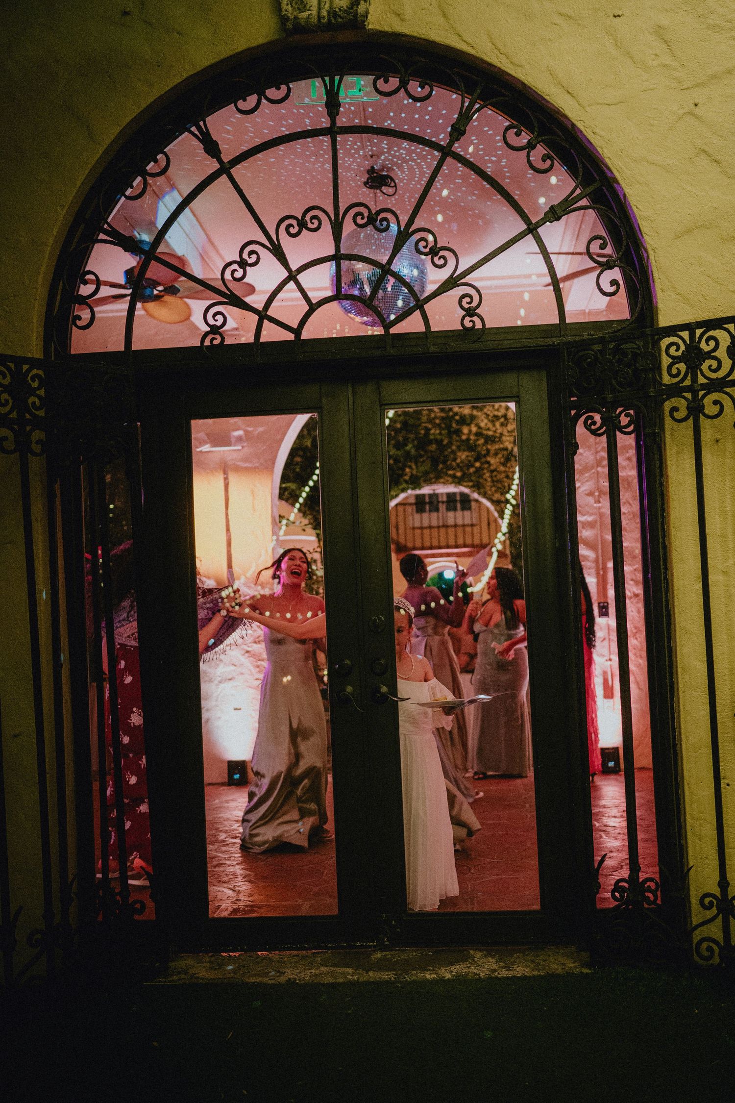Elegant French doors with ornate ironwork reveal a wedding reception inside, glowing with warm pink lights.