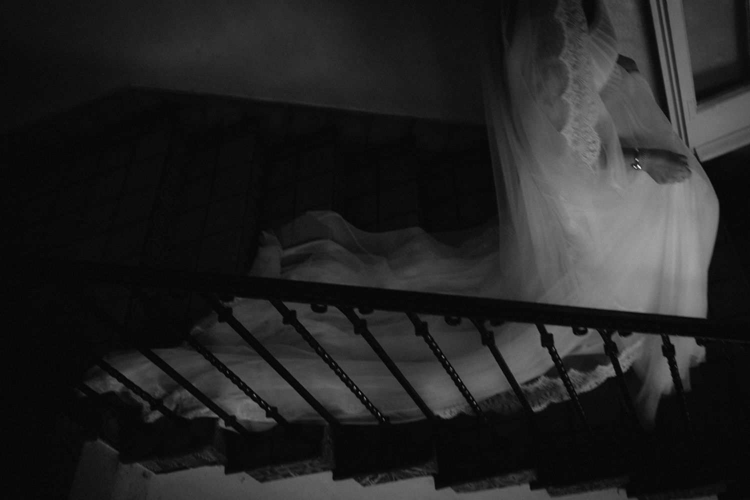 Ethereal bride in white wedding dress descending a dark staircase, captured in dramatic black and white photography.