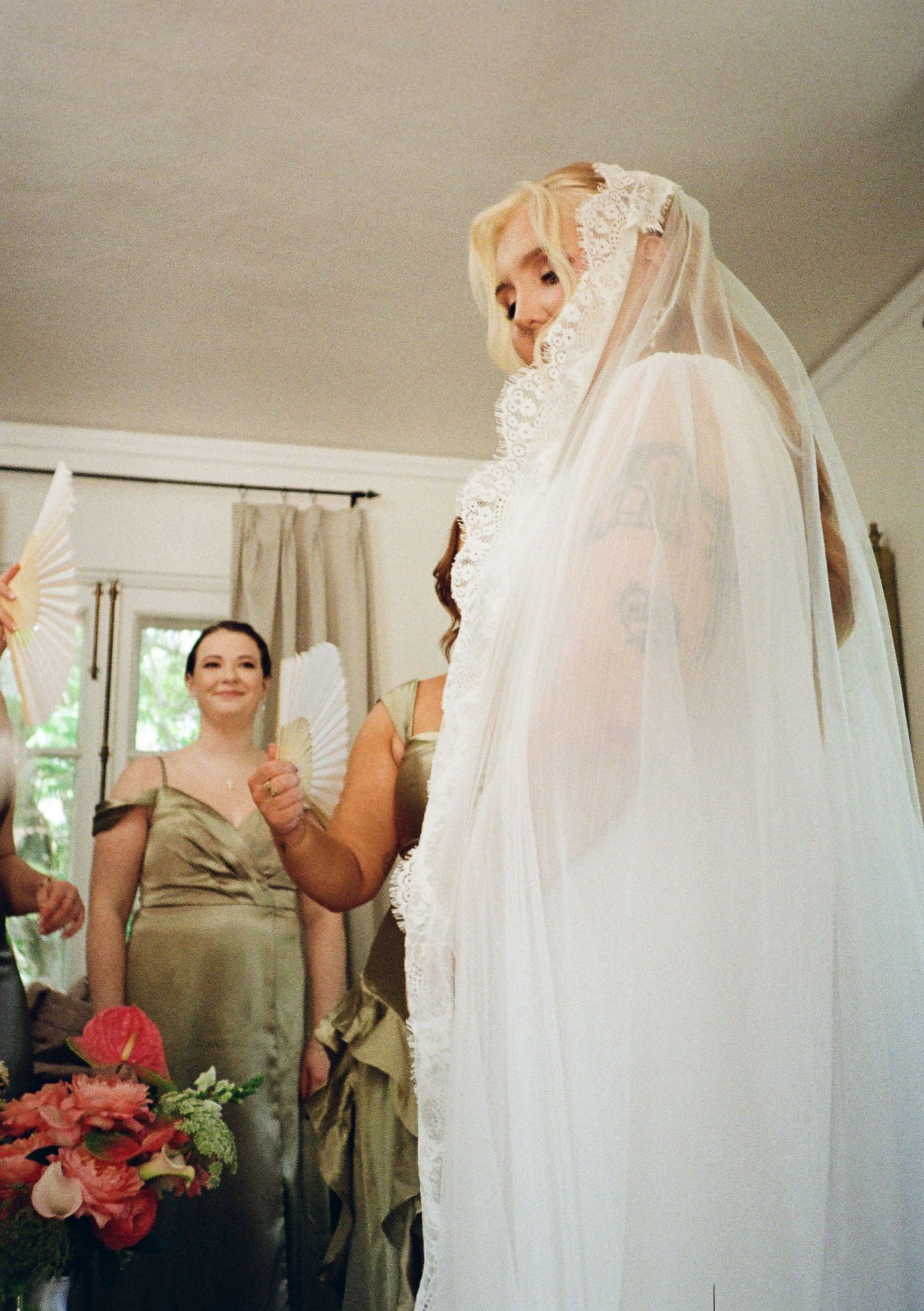Bride in lace veil with bridesmaid in olive gown holding pink flowers indoors.