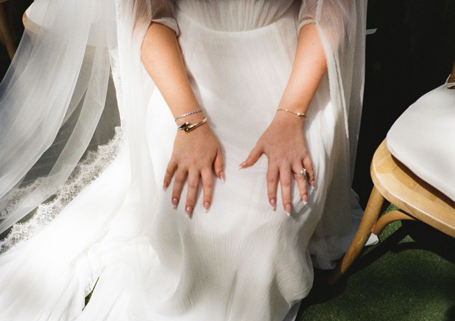Bride in white wedding dress with gold bracelets and rings, hands resting on flowing gown skirt.