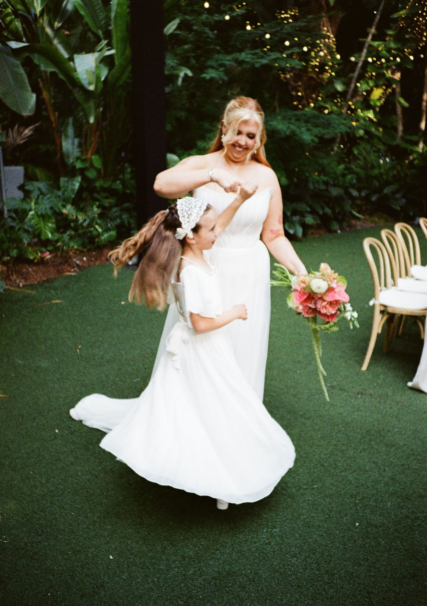 Bride in white gown twirling flower girl in matching white dress outdoors at garden wedding ceremony.