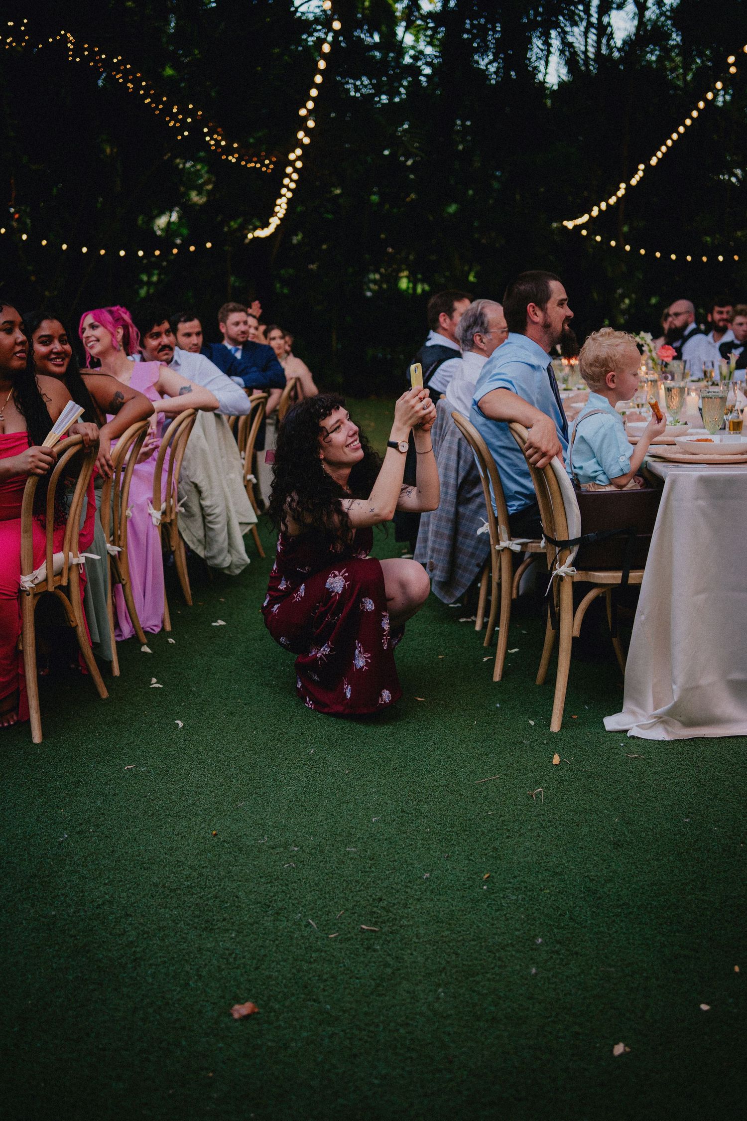 Outdoor wedding reception on grass with string lights, guests seated at tables while a woman in burgundy dress sits on the lawn.