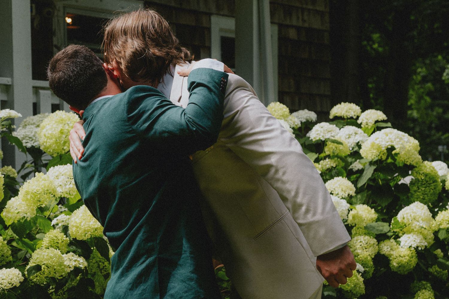 Two people share a warm embrace in front of blooming white hydrangeas outside a home.