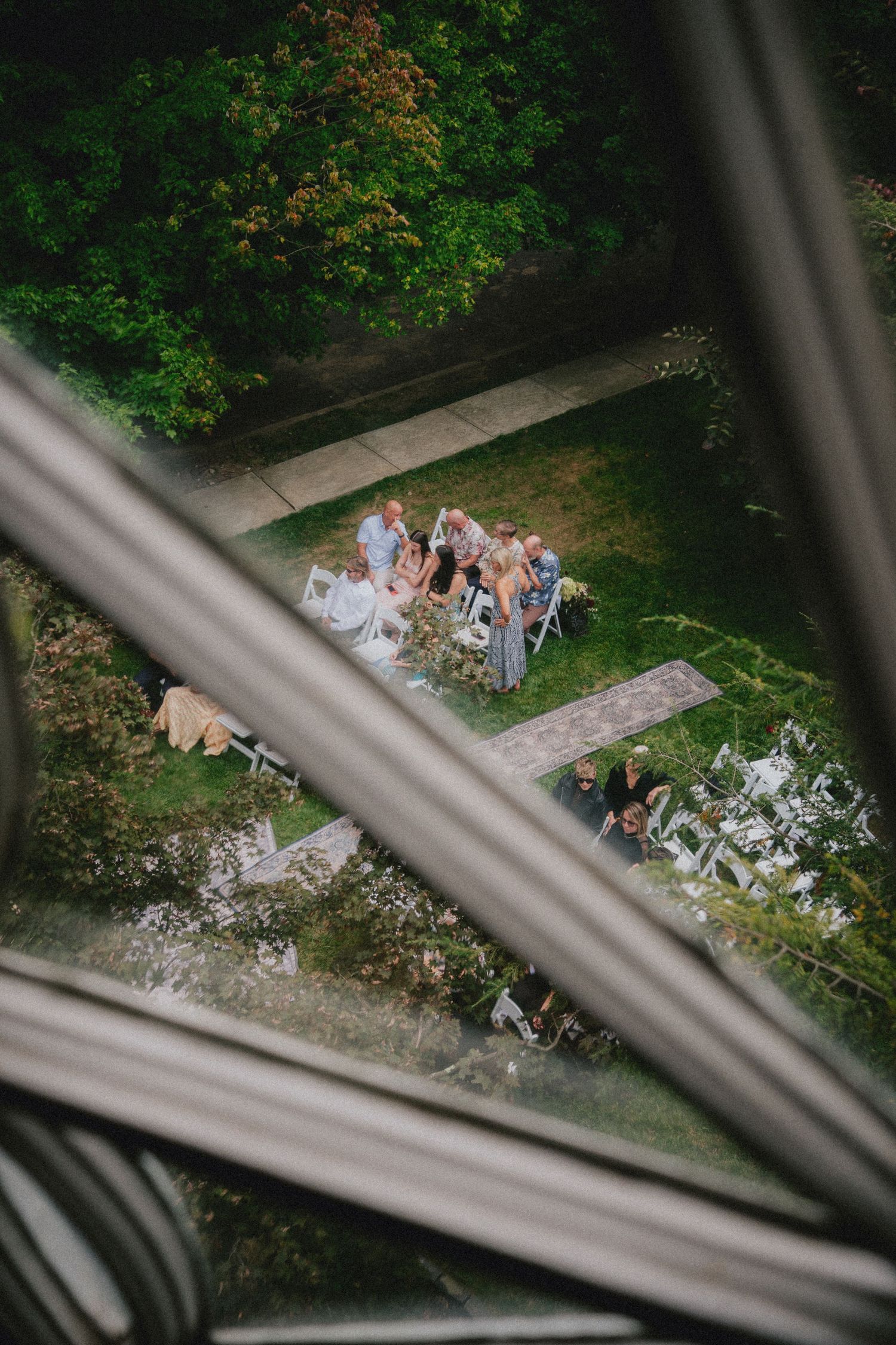 Aerial view of an outdoor wedding ceremony on a green lawn, with guests seated in white chairs surrounded by lush trees.