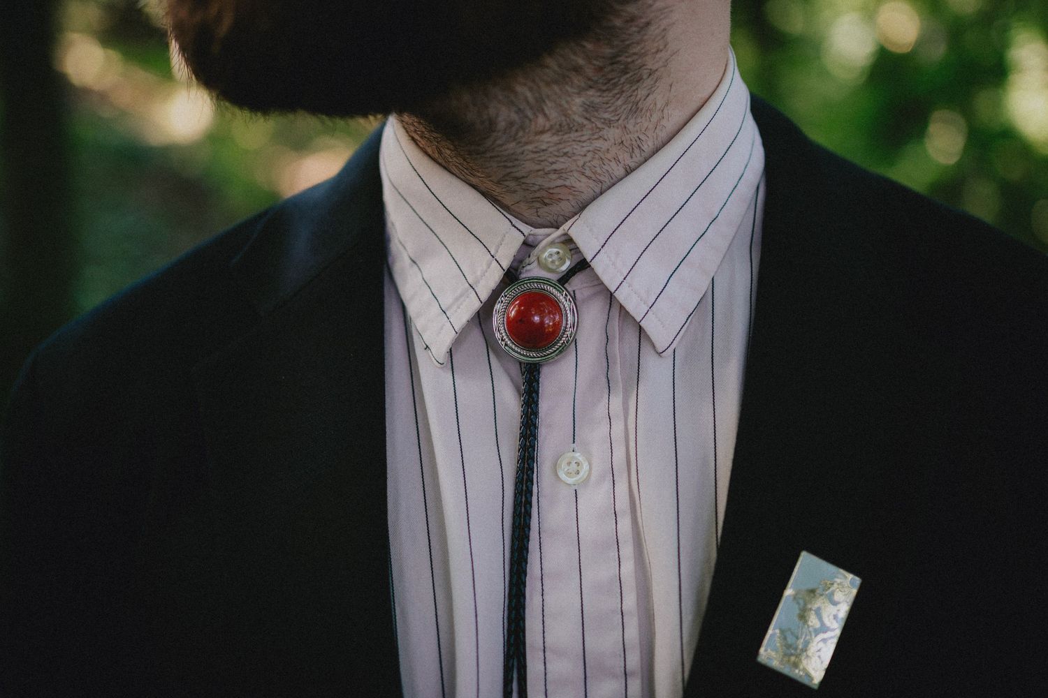 Man in black suit wearing a bolo tie with red stone centerpiece and boutonniere on striped dress shirt.