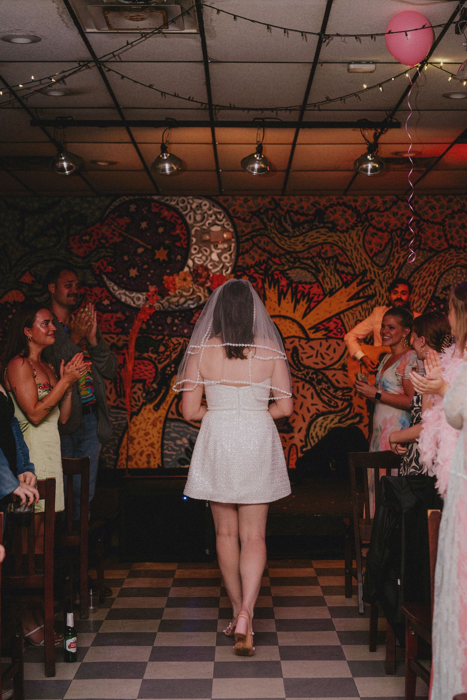 Bride in short white dress and veil walks down aisle in decorated venue with colorful mural wall.