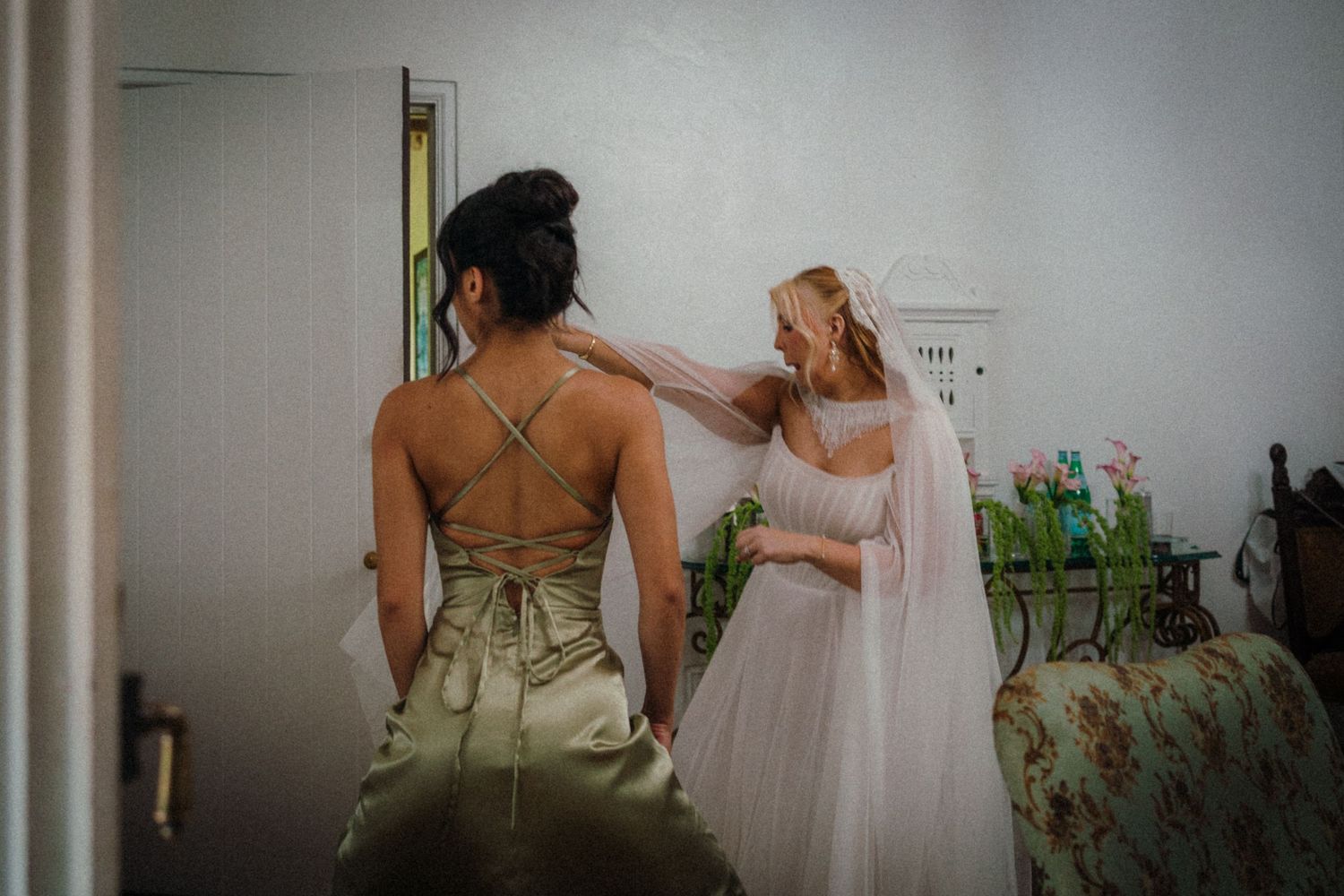 Bride in white gown and veil being assisted by bridesmaid in satin dress during wedding preparations.
