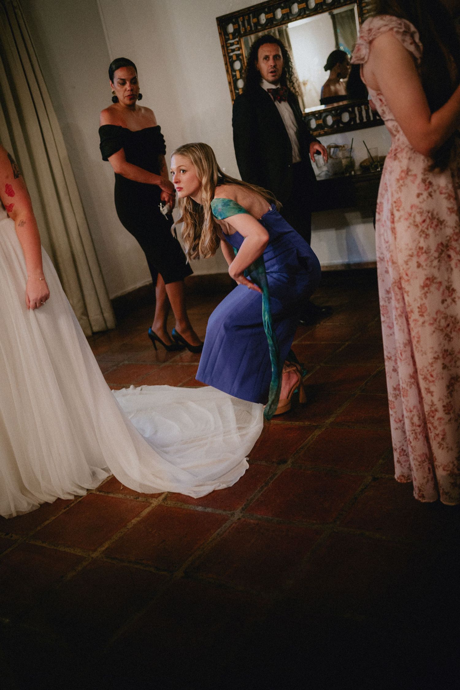 Bridesmaid in blue dress adjusting bridal gown train while guests look on in elegant venue.