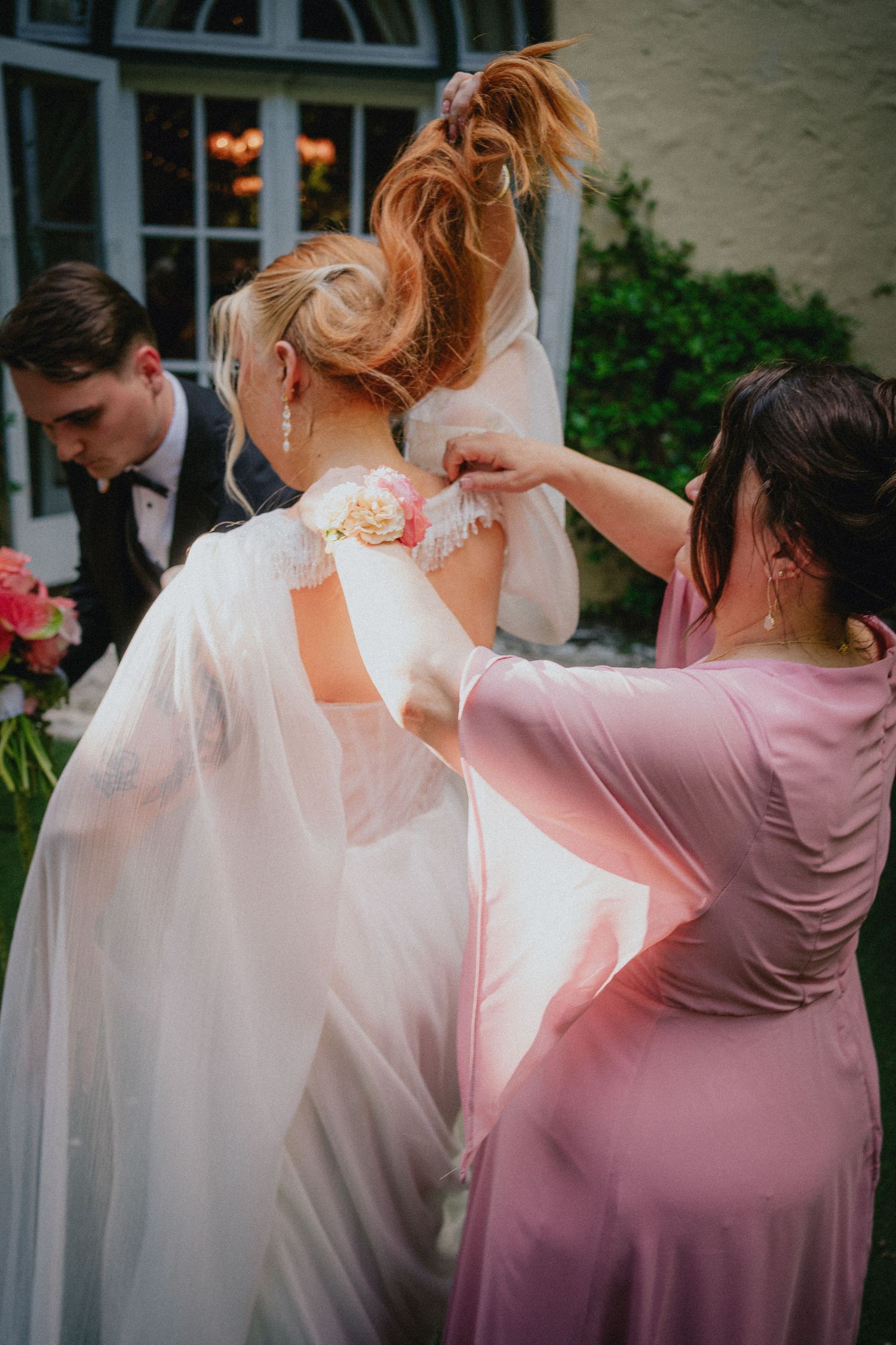 Bridesmaids helping bride with her wedding dress outdoors, adjusting lace details on elegant white bridal gown.