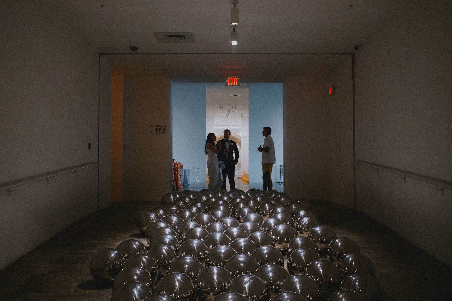 Art installation with reflective silver spheres on gallery floor, three visitors standing in softly lit museum space.