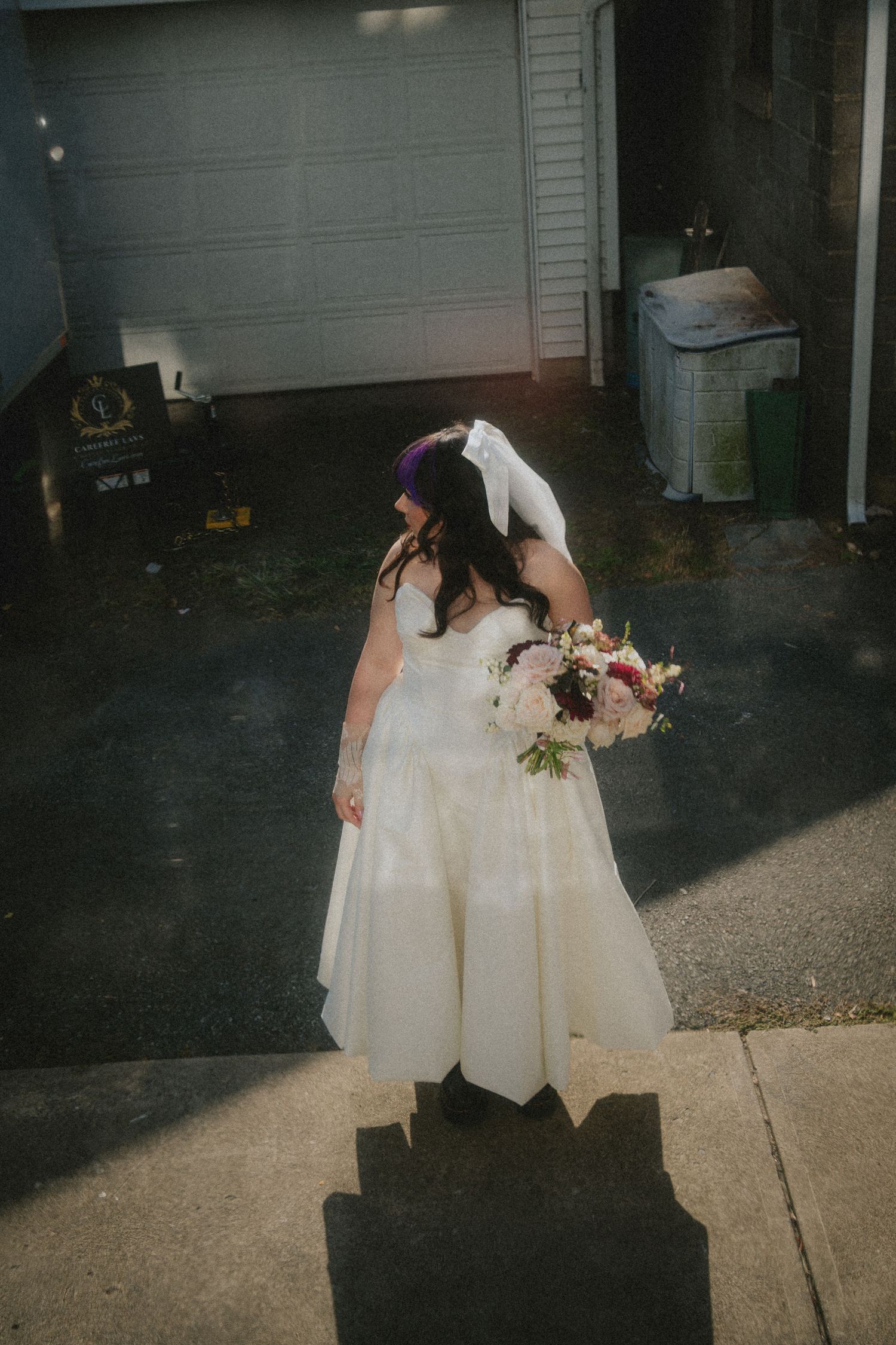 Bride in white wedding gown holding colorful bouquet stands near garage, veil lifted by breeze on wedding day.
