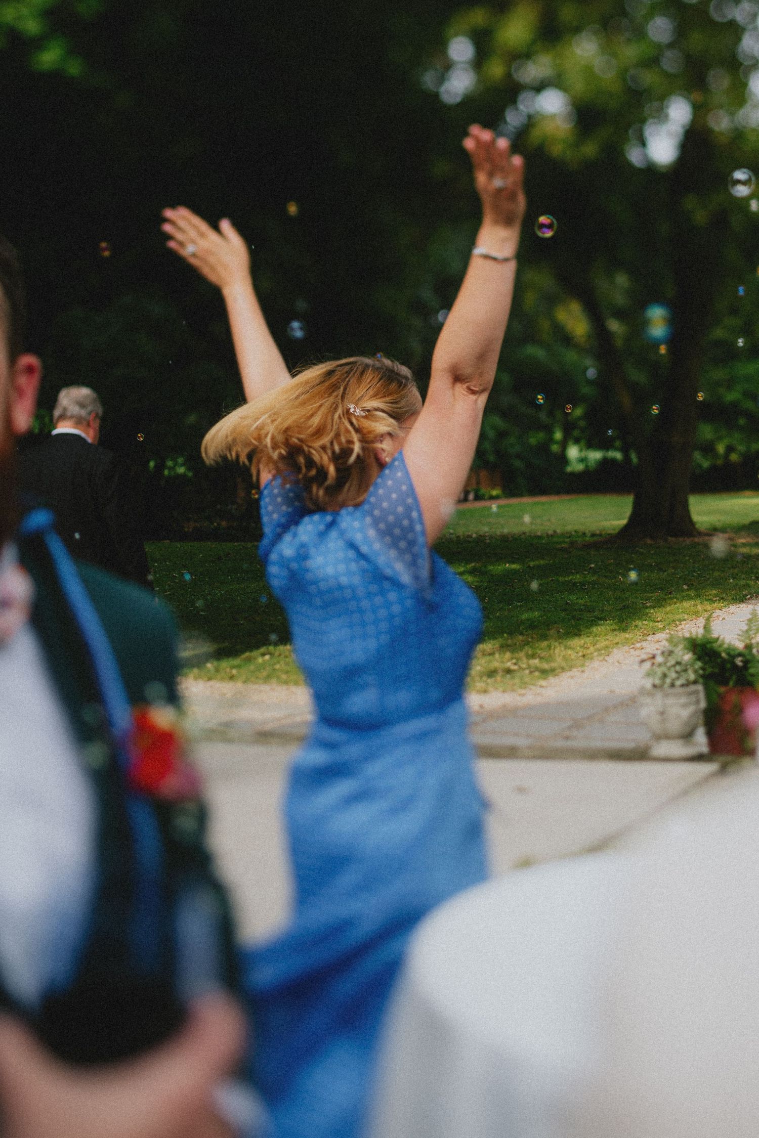 Woman in blue polka dot dress raises arms joyfully outdoors at a wedding celebration surrounded by guests.