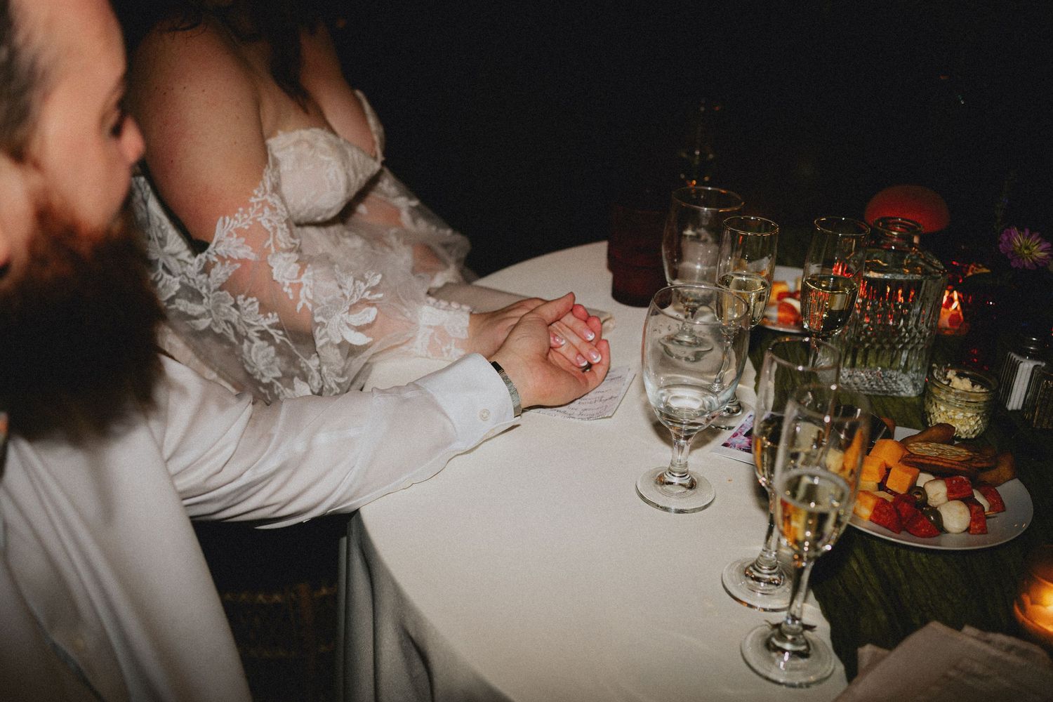 Bride and groom holding hands at wedding reception table with elegant glassware and floral centerpiece.