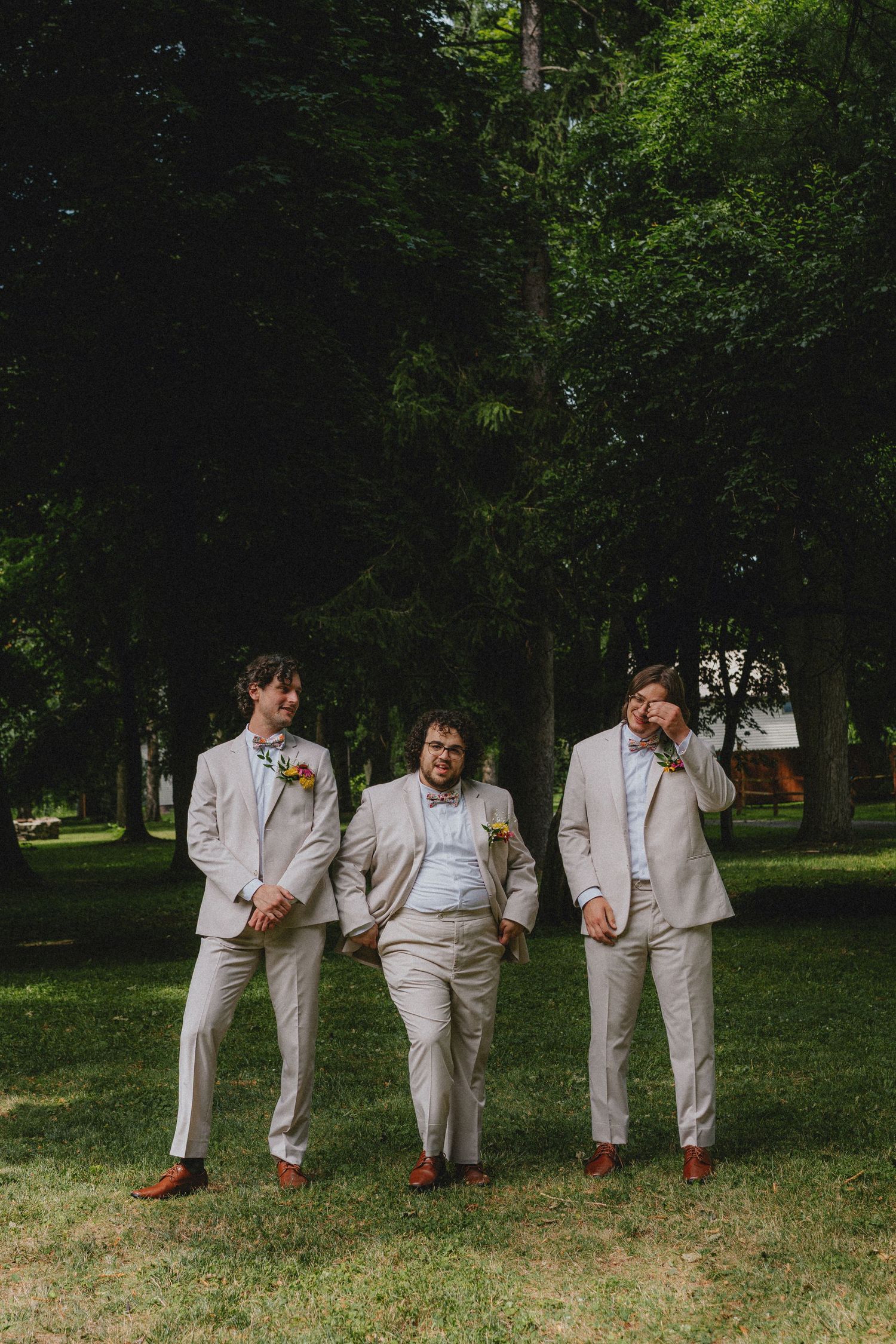 Three groomsmen in matching tan suits with boutonnieres pose outdoors on green grass with tall trees in background.