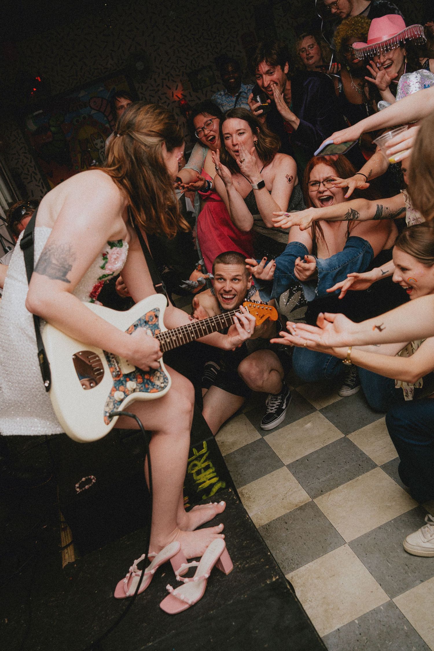 Guitarist performs seated on stage edge while excited crowd reaches out at an intimate indoor concert venue.