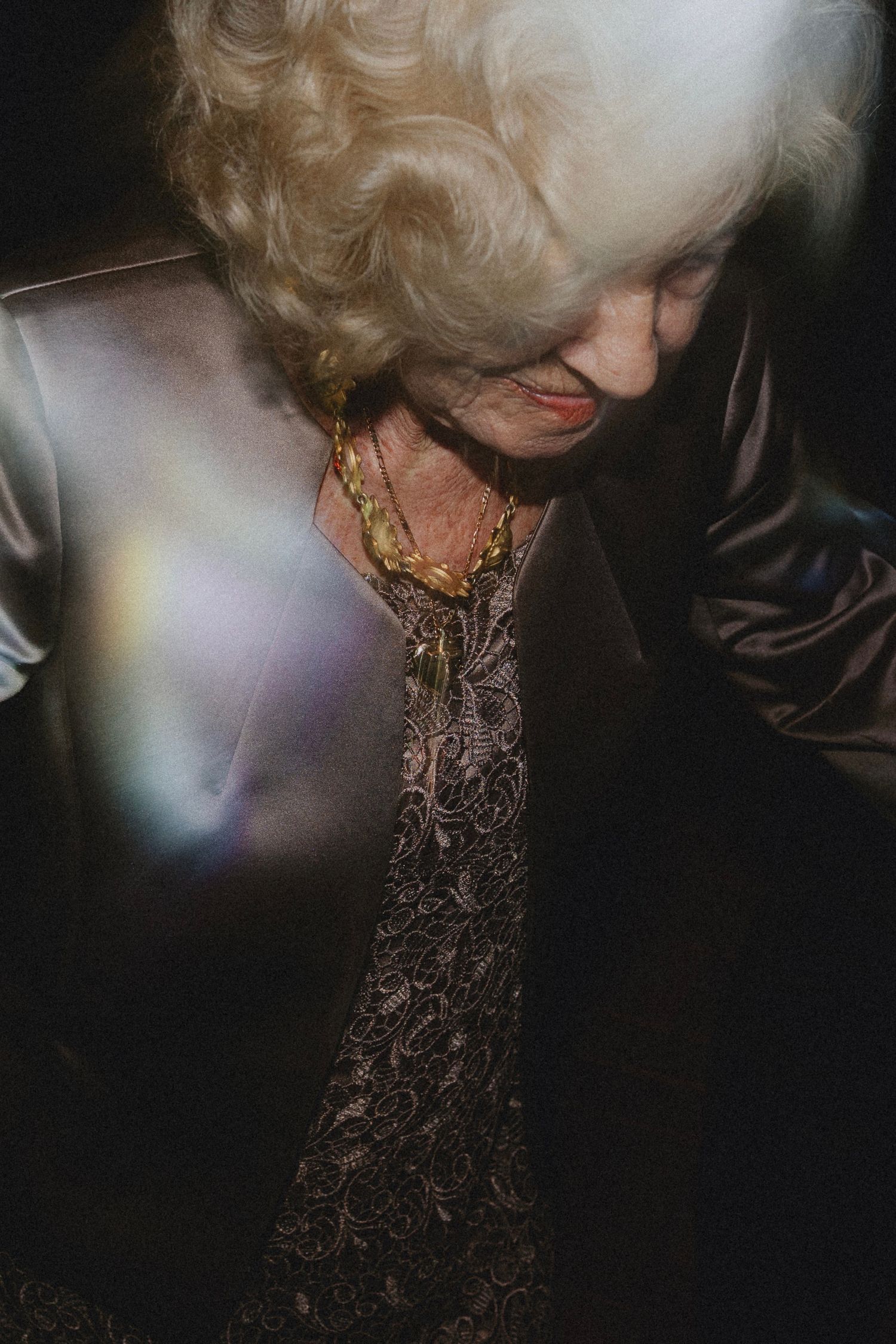 Elderly woman with white curly hair wearing lace dress and gold necklace, on the dance floor with rainbow light flare.