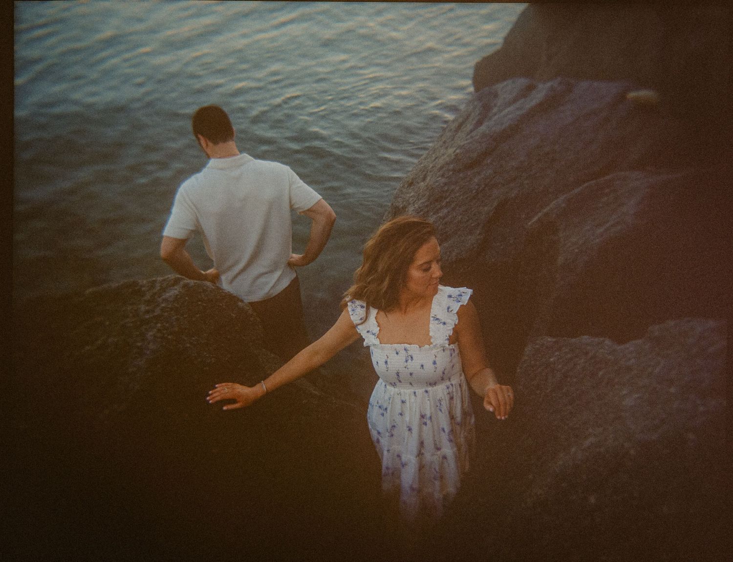 Couple near rocky shoreline, woman in white floral dress and man in white shirt standing by water at sunset.