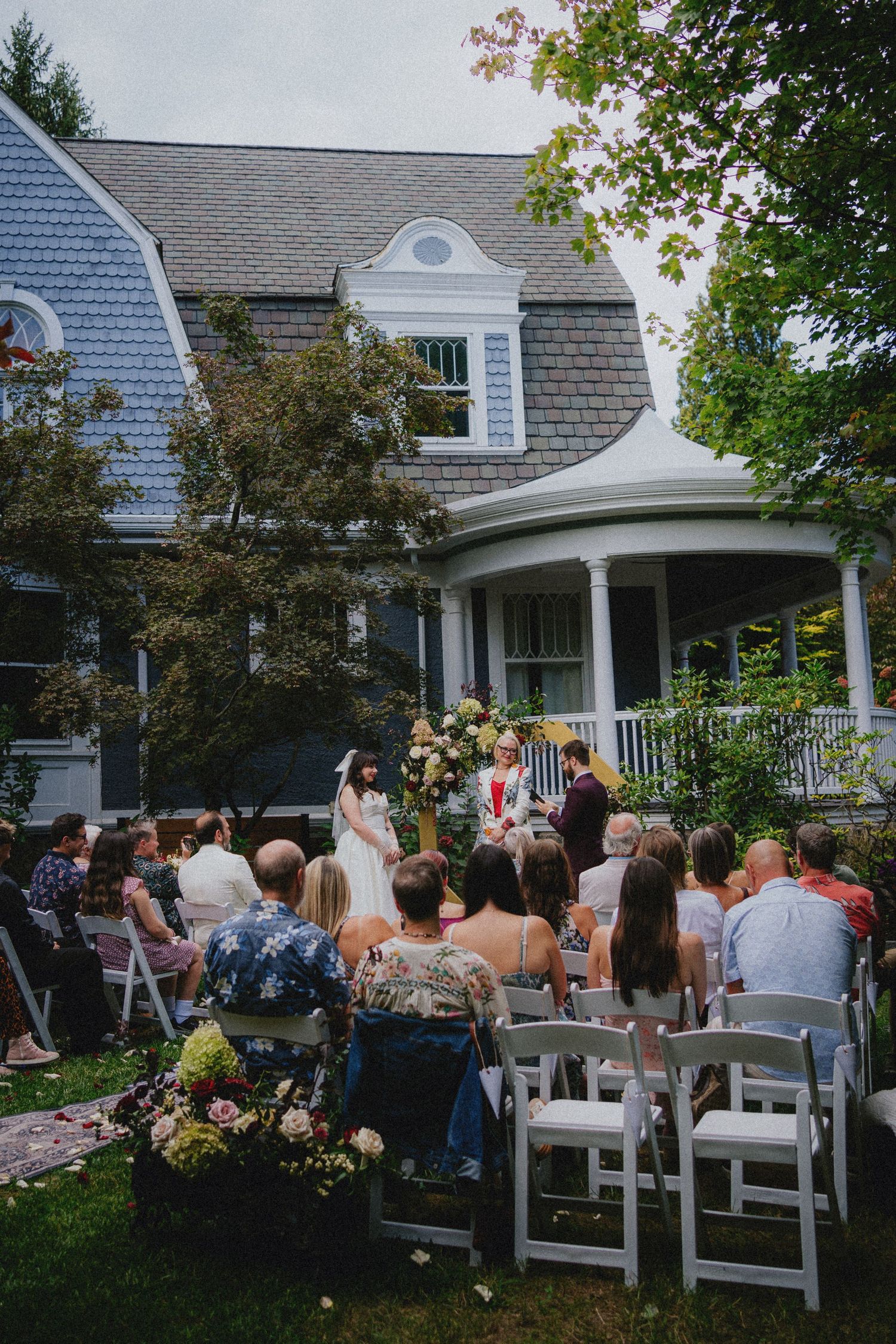 Outdoor wedding ceremony in a lush backyard with white chairs, guests seated, and a charming blue shingled house backdrop.