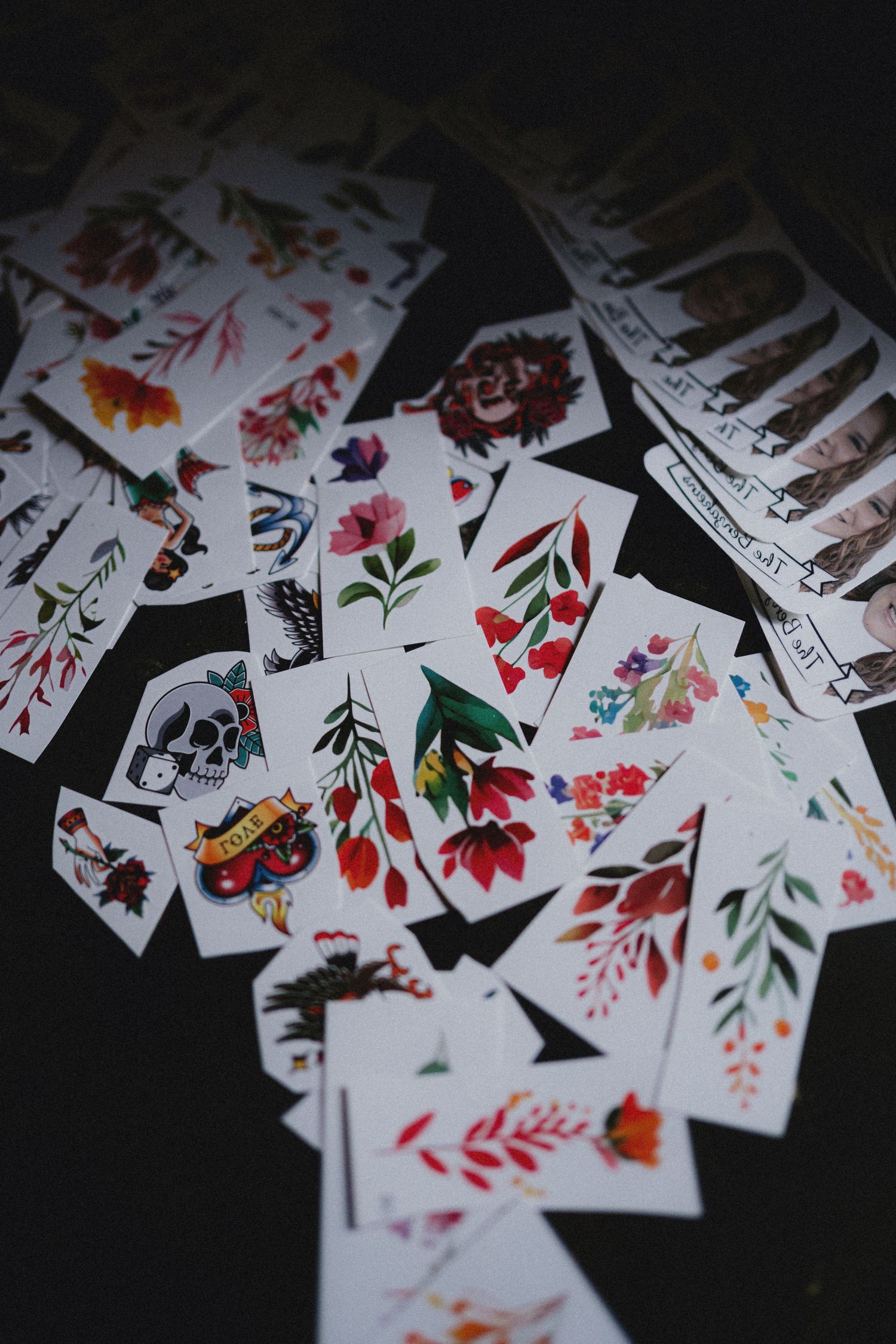 Colorful floral and skull temporary tattoo flash cards spread across a dark surface.