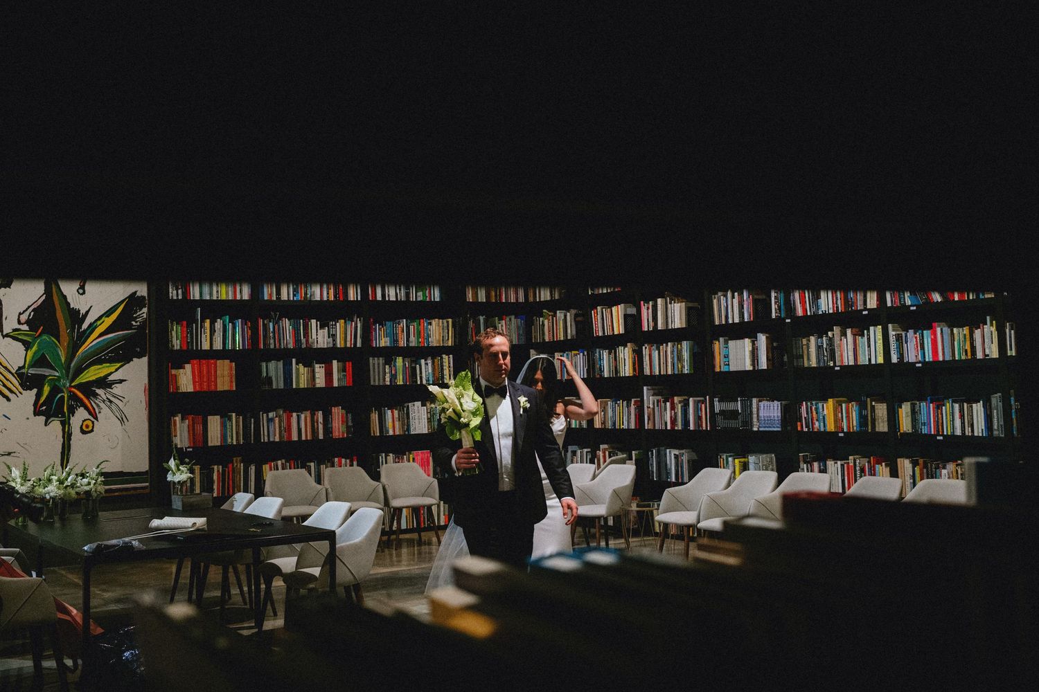 Person standing in a dimly lit library with floor-to-ceiling bookshelves, holding flowers during an event.
