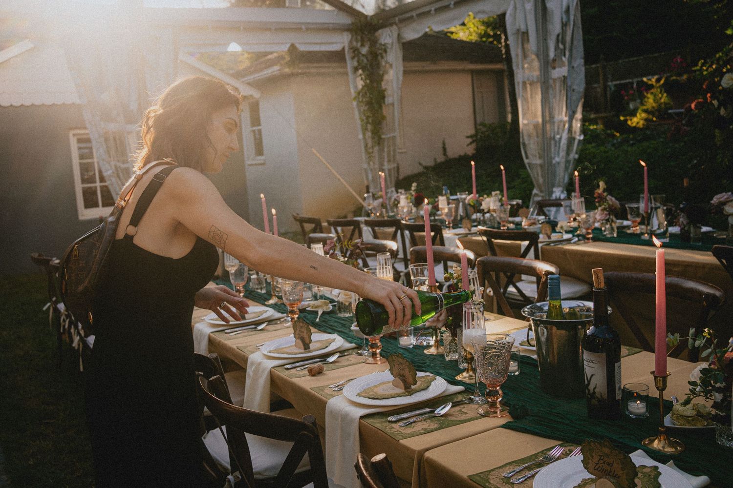 Woman in black dress pouring wine at elegantly set outdoor dinner table with candles at golden hour.