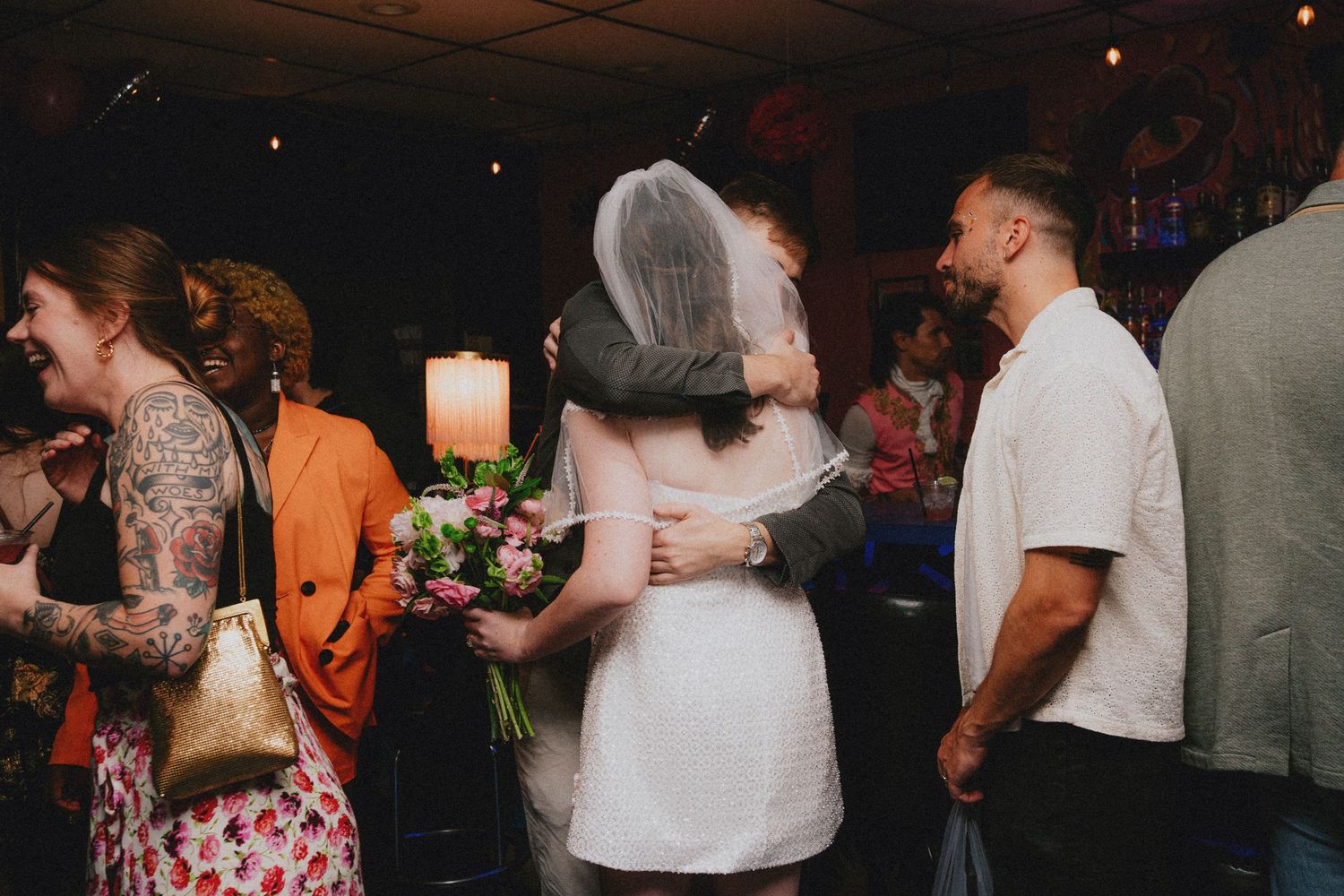 Bride in white dress and veil hugging guest at wedding reception, surrounded by colorful party guests.