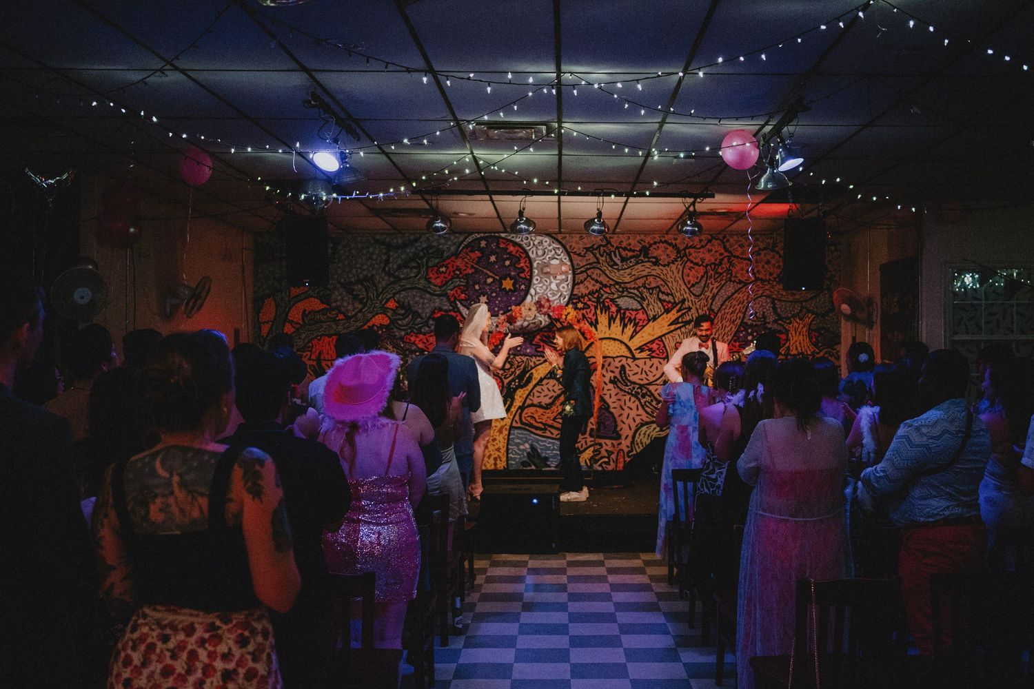 Band performing live on a colorful mural-adorned stage in a dimly lit venue with string lights and a dancing crowd.