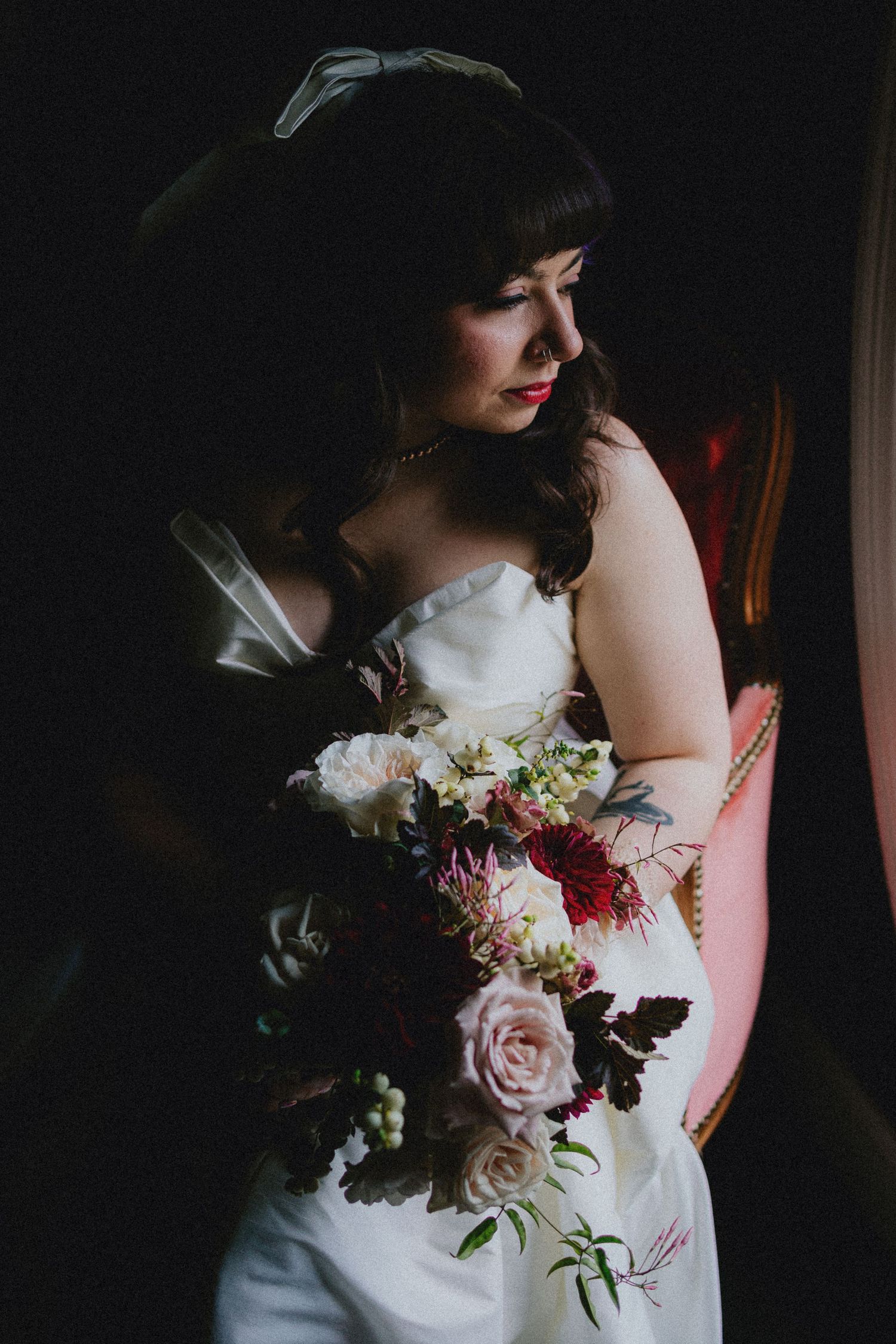Bride in strapless white gown holds lush bouquet of pink roses and burgundy blooms in dramatic moody lighting.