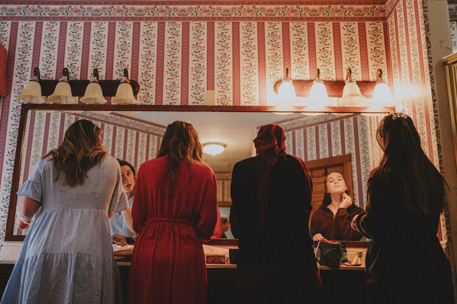 Four women stand before a vintage mirror in a floral wallpapered room with warm wall sconce lighting.