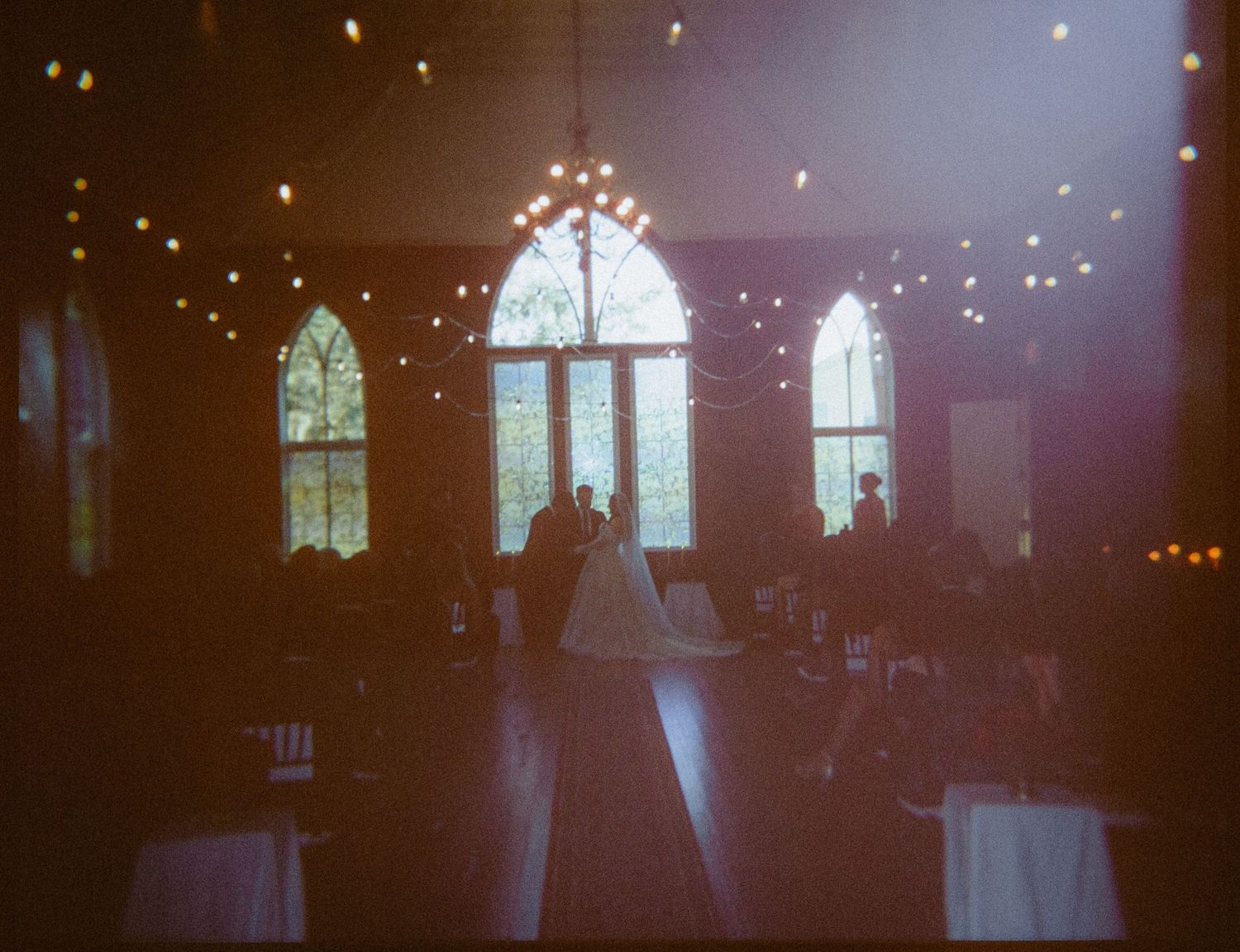 Couple sharing first dance in elegant wedding venue with gothic arched windows and warm string lights overhead.