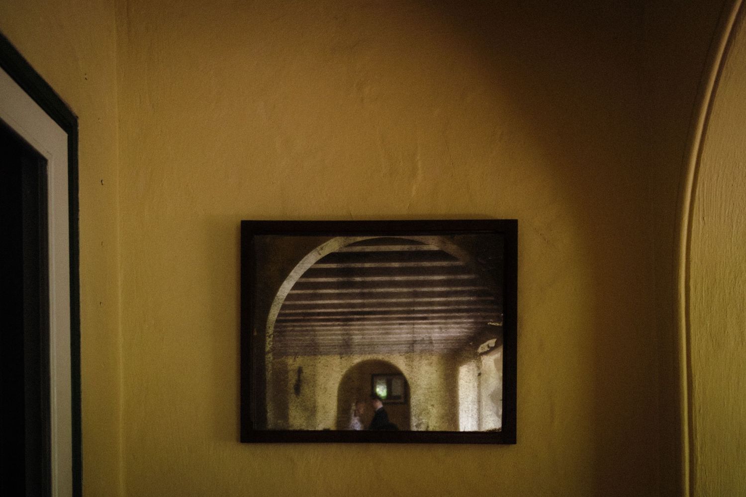 Framed photograph of an arched corridor hanging on a dimly lit yellow wall beside a dark doorframe.