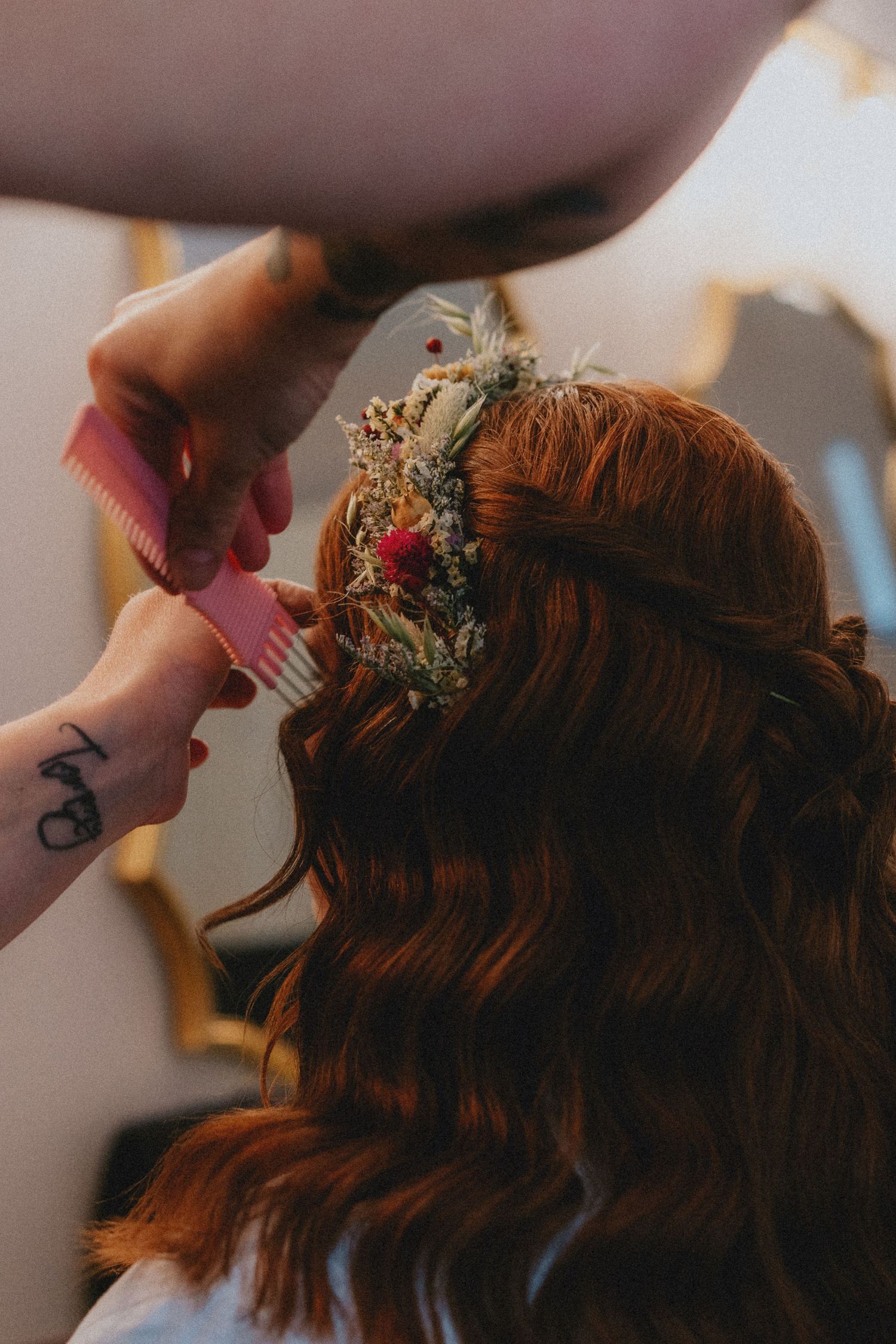 Hairstylist using a pink comb to style a bride's auburn hair adorned with a floral crown of dried flowers.