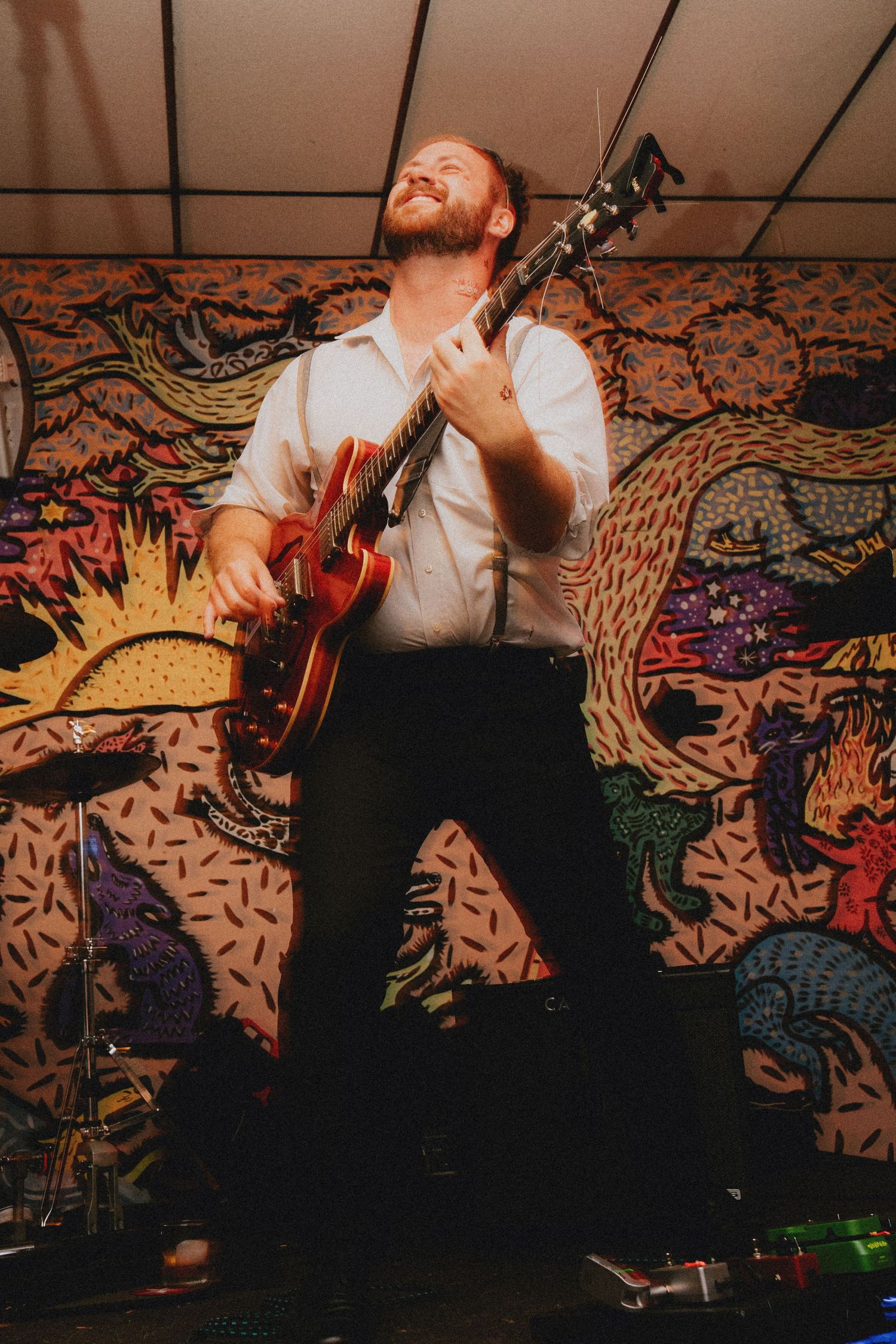 Guitarist performing on stage with colorful mural backdrop, wearing white shirt, playing red electric guitar with joy.