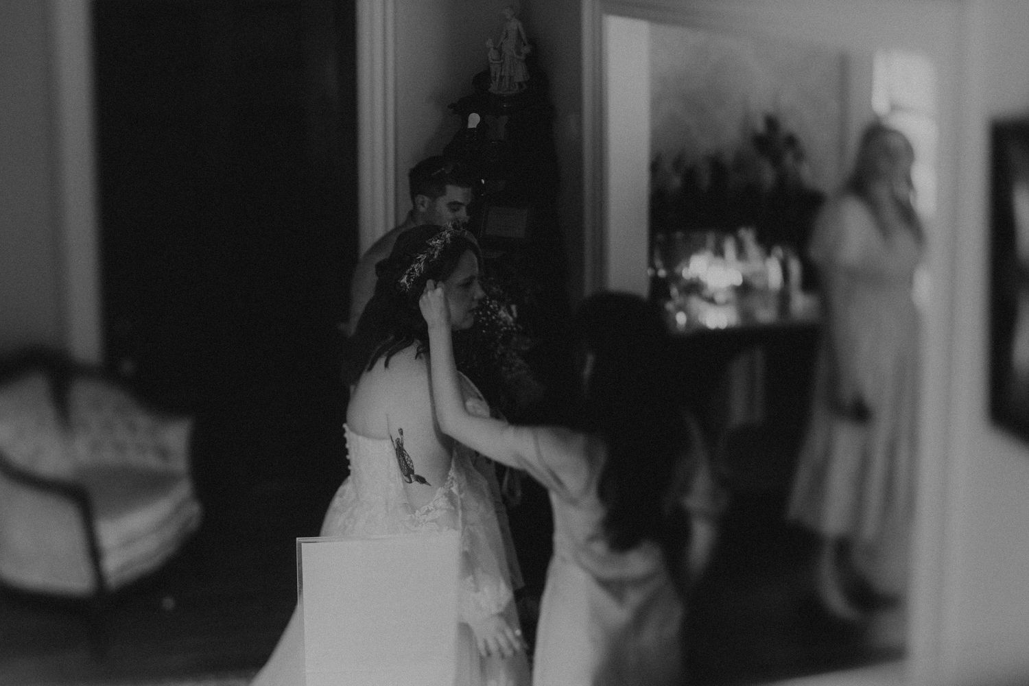 Black and white wedding photo of bride getting ready with bridesmaids helping in elegant interior.