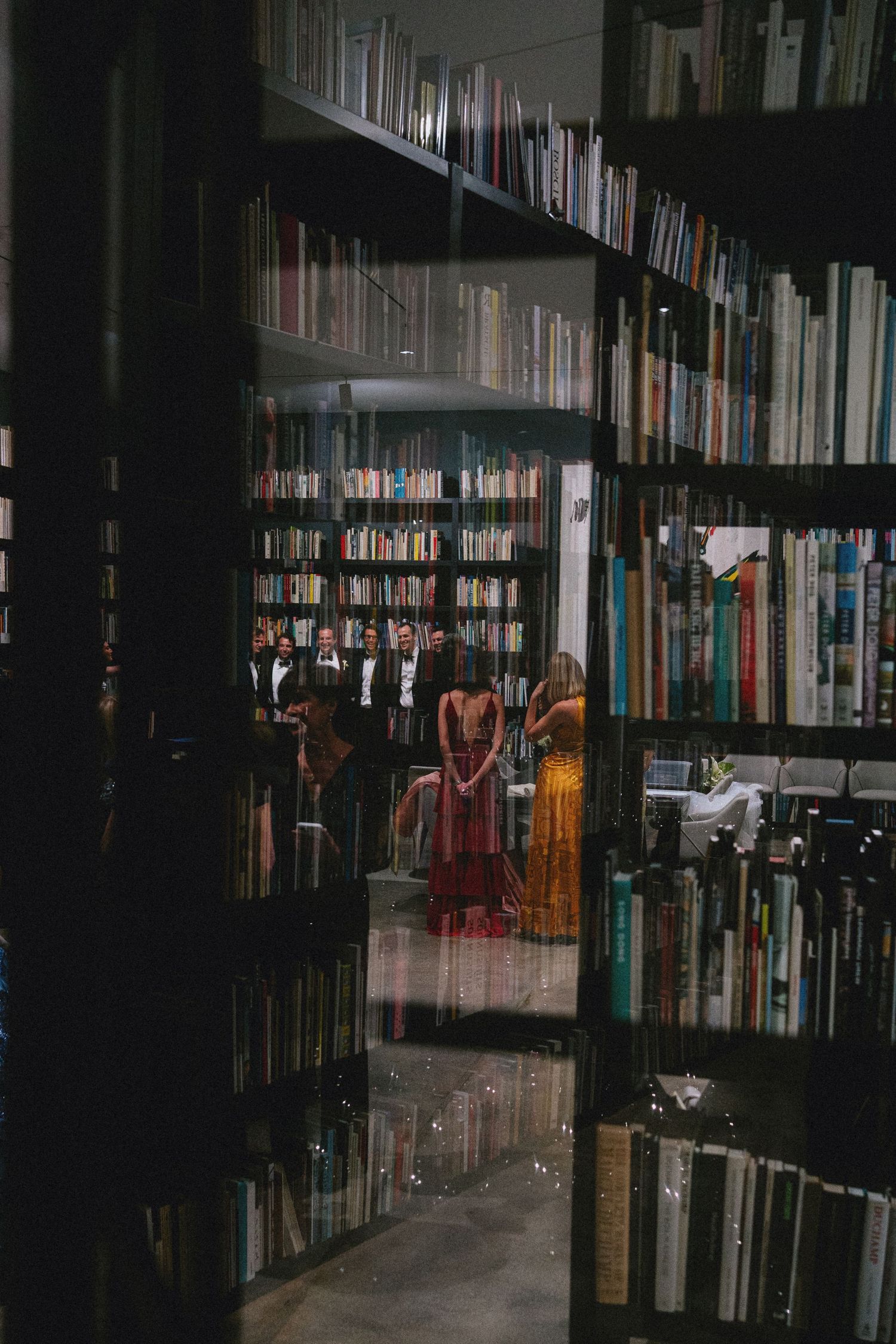 People in formal attire gathered in a dimly lit library with floor-to-ceiling bookshelves, viewed through glass doors.