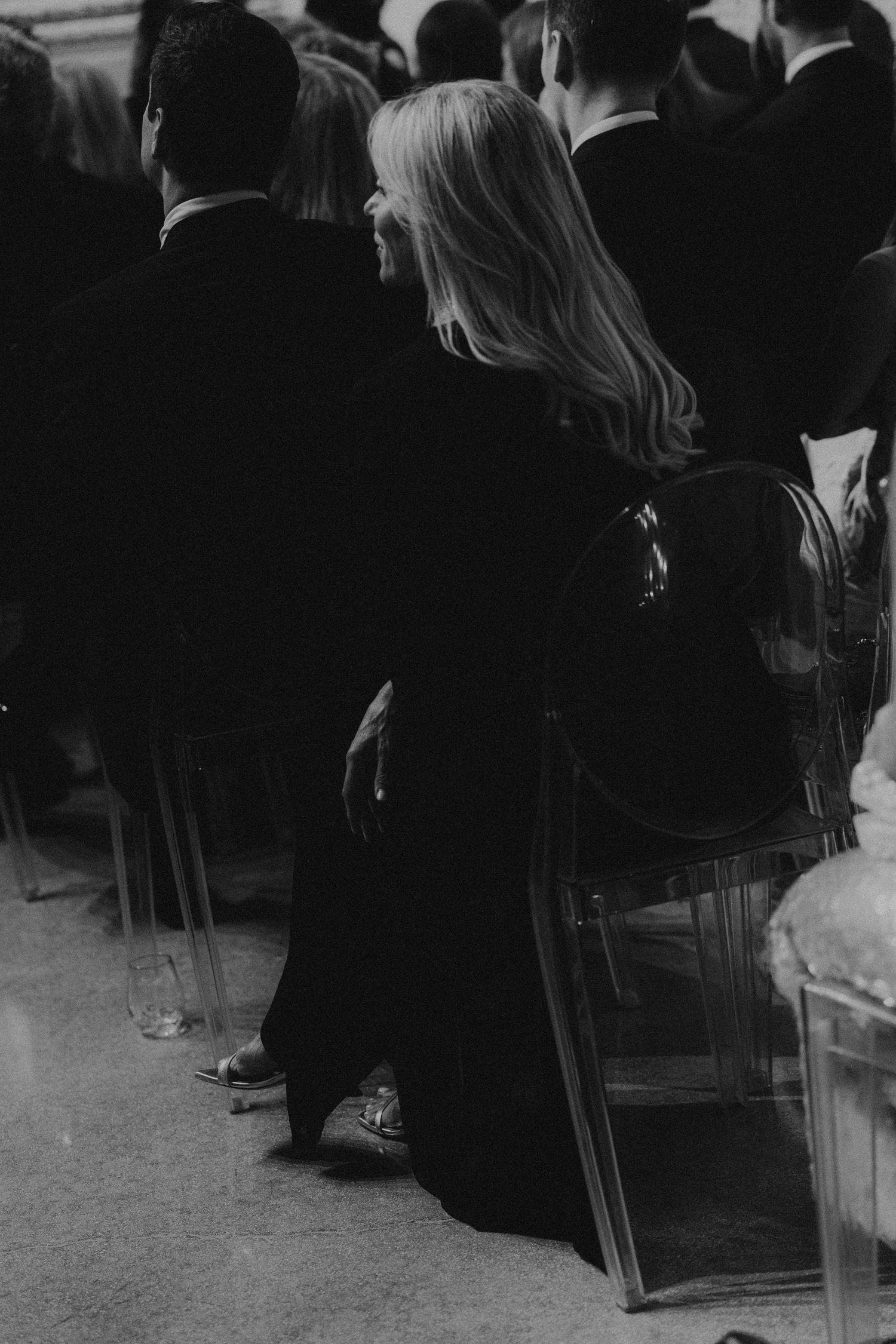 Black and white photo of a blonde woman in a dark gown seated in a clear chair at a formal event.