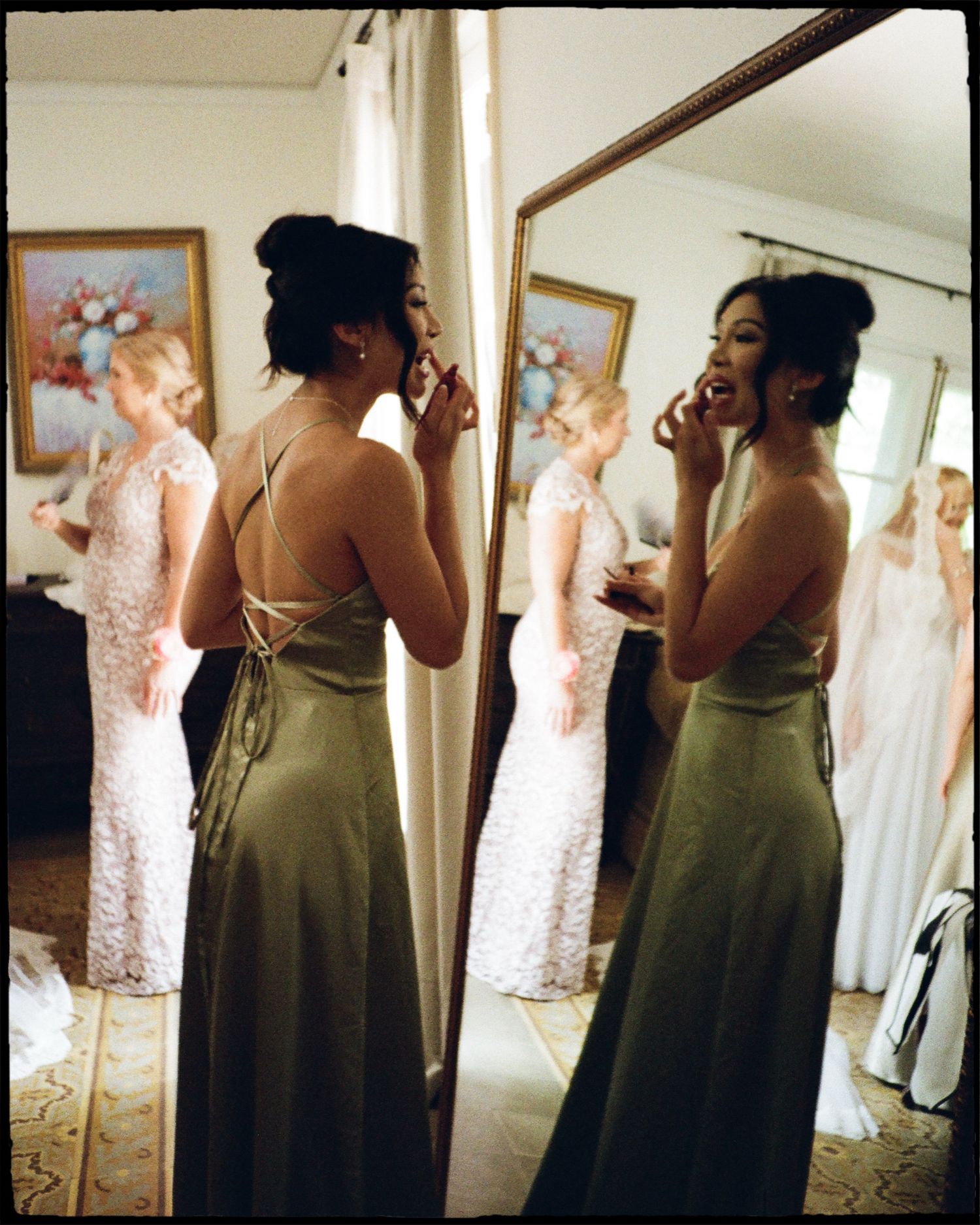 Bridesmaid in olive green satin gown applying makeup before mirror, with bride in white lace dress visible in reflection.