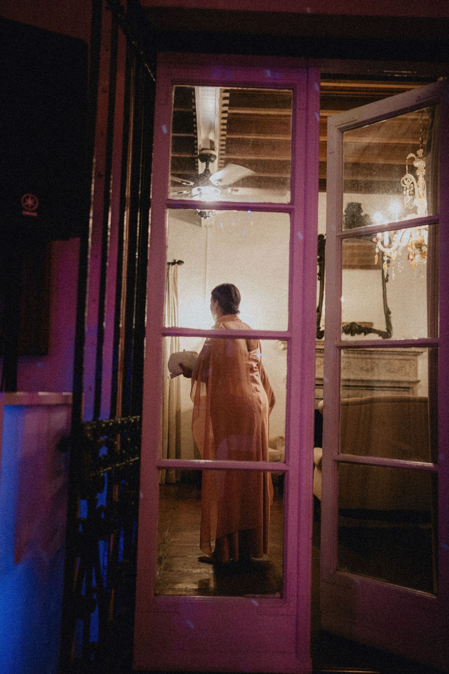 Person in flowing gown viewed through pink French doors in dimly lit elegant room with chandelier.