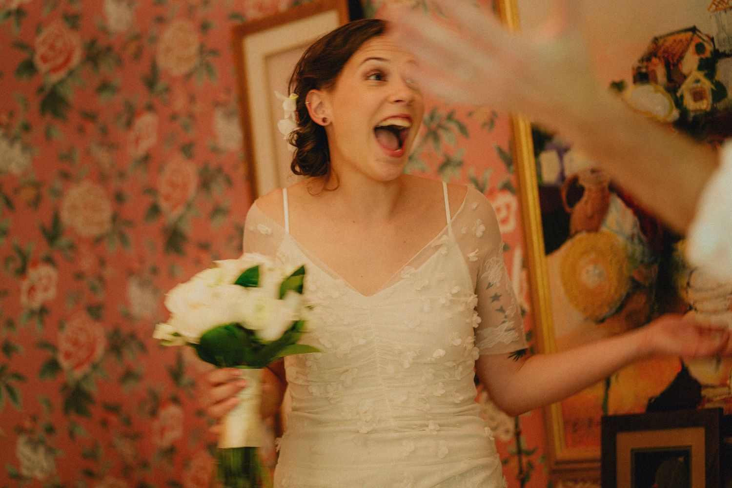 Bride in white wedding dress laughing joyfully while holding white rose bouquet in floral wallpaper room.