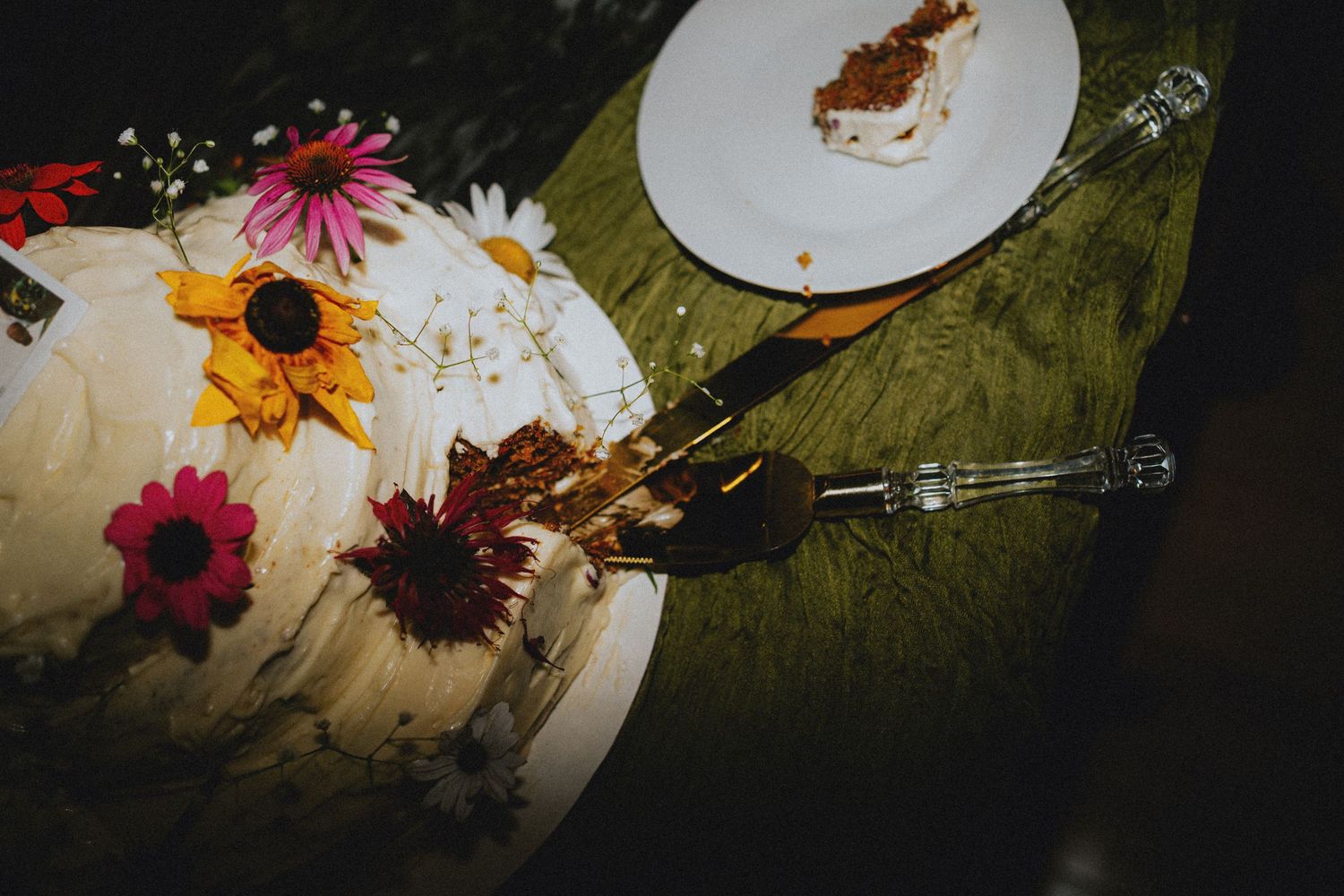 White wedding cake decorated with colorful wildflowers including sunflowers and daisies, with a slice served on a plate nearby.