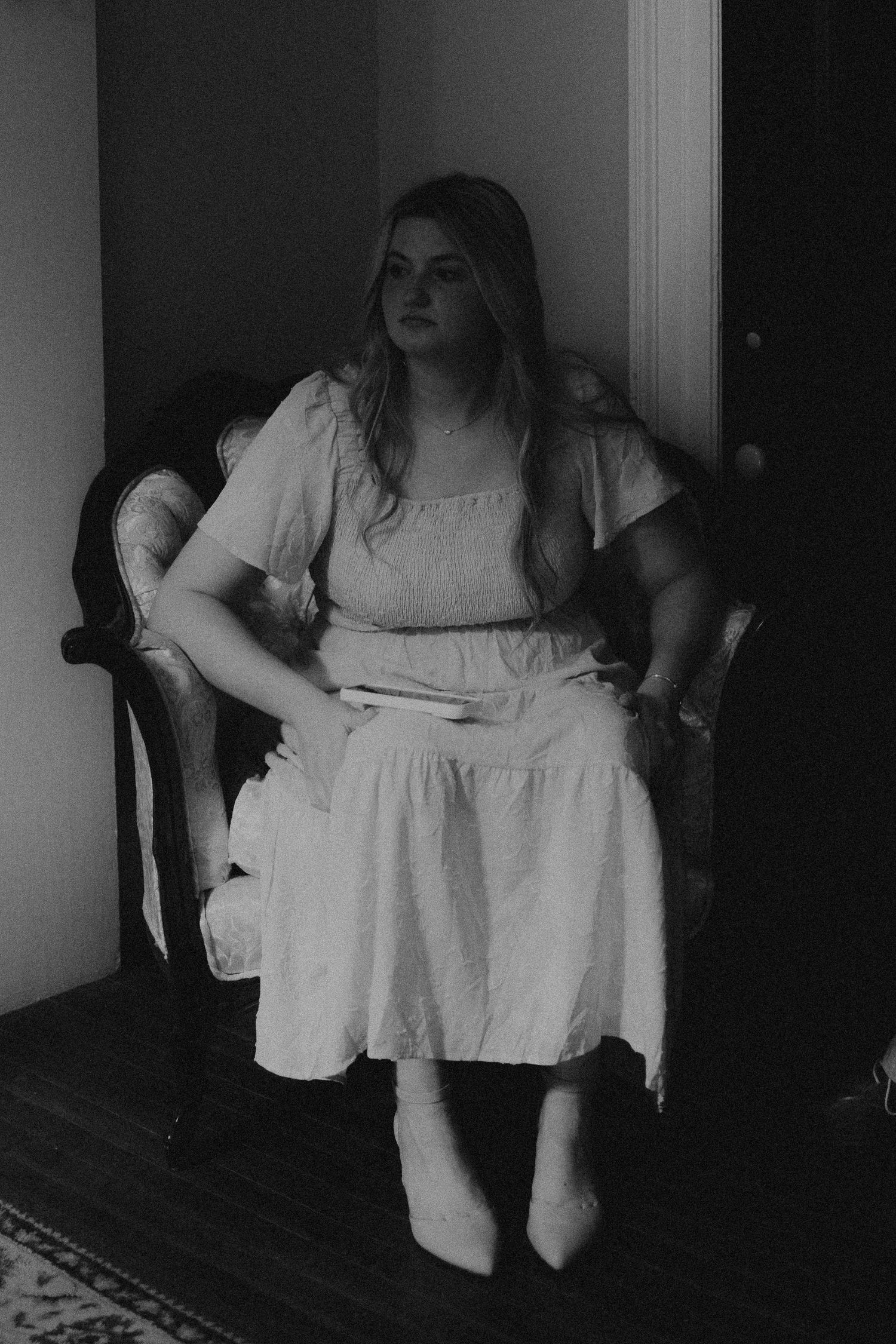 Black and white photo of a young woman in a white dress sitting in an ornate chair in a dimly lit room.