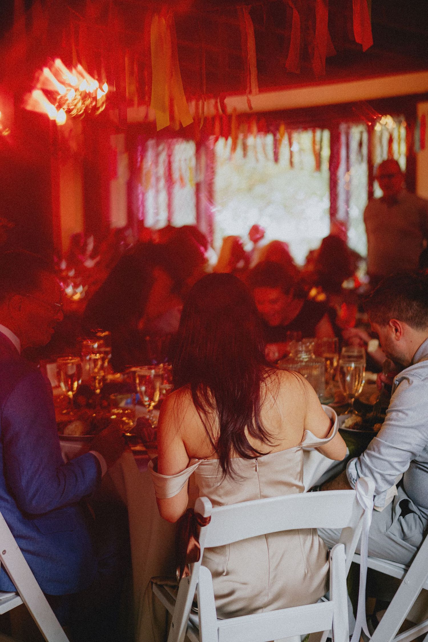 Guests seated at elegantly decorated wedding reception tables under warm red lighting with colorful fringe decorations.